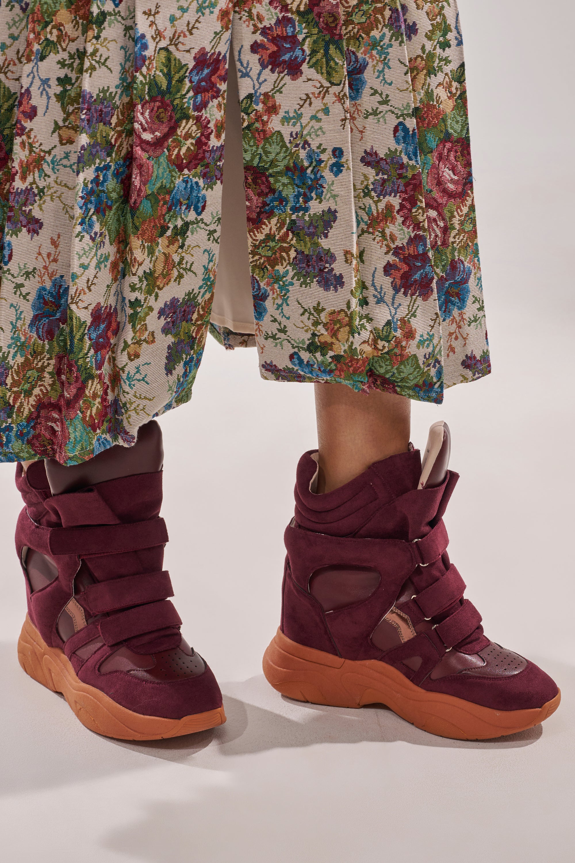 A person wearing a long, floral-patterned skirt and MARANTO BURGUNDY WEDGE FAUX SUEDE SNEAKERS with thick soles and wide straps stands on a light-colored surface. 1