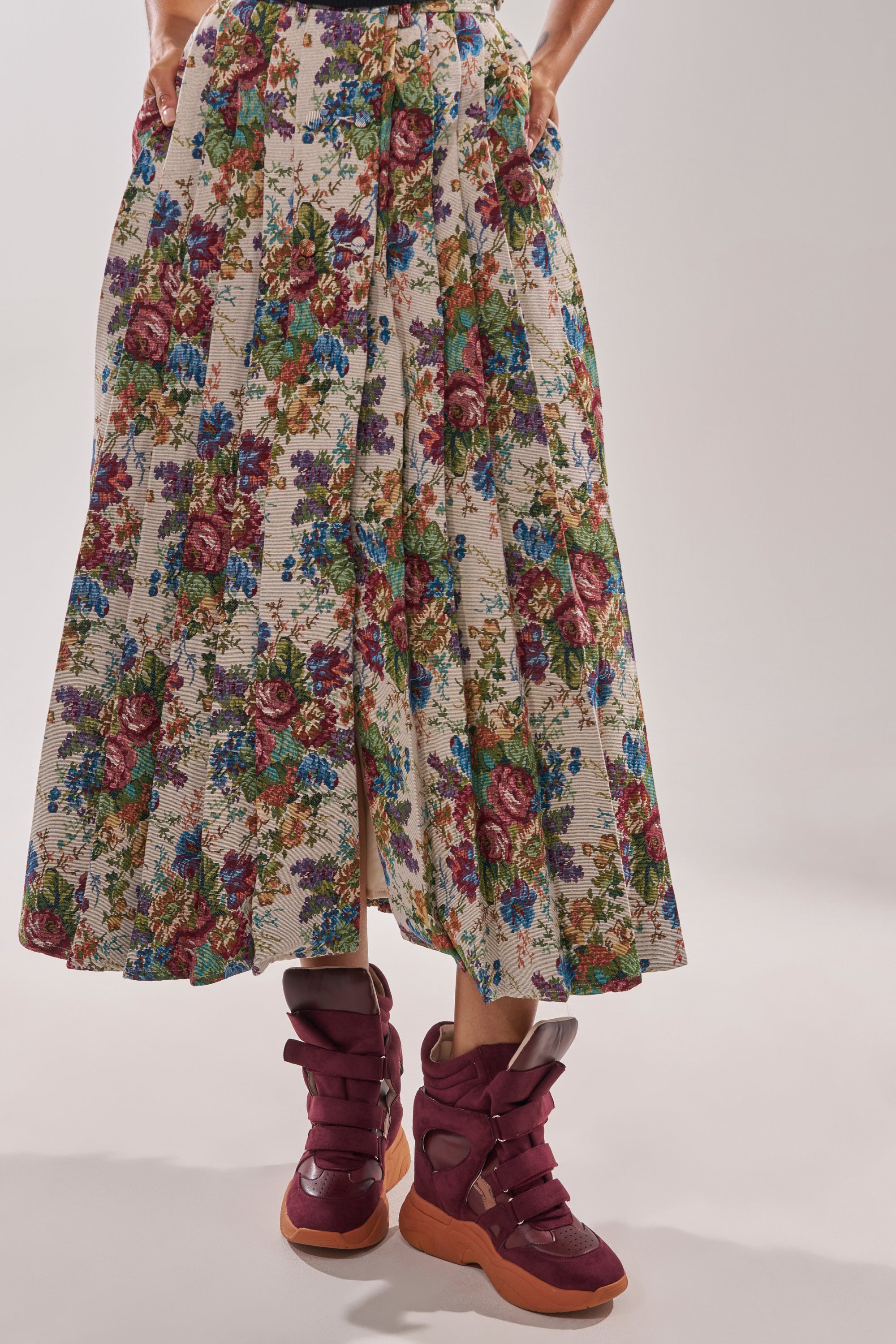 Wearing a long, floral-patterned skirt with hands in pockets and MARANTO BURGUNDY WEDGE FAUX SUEDE SNEAKER, a person stands against a plain, light background. 2