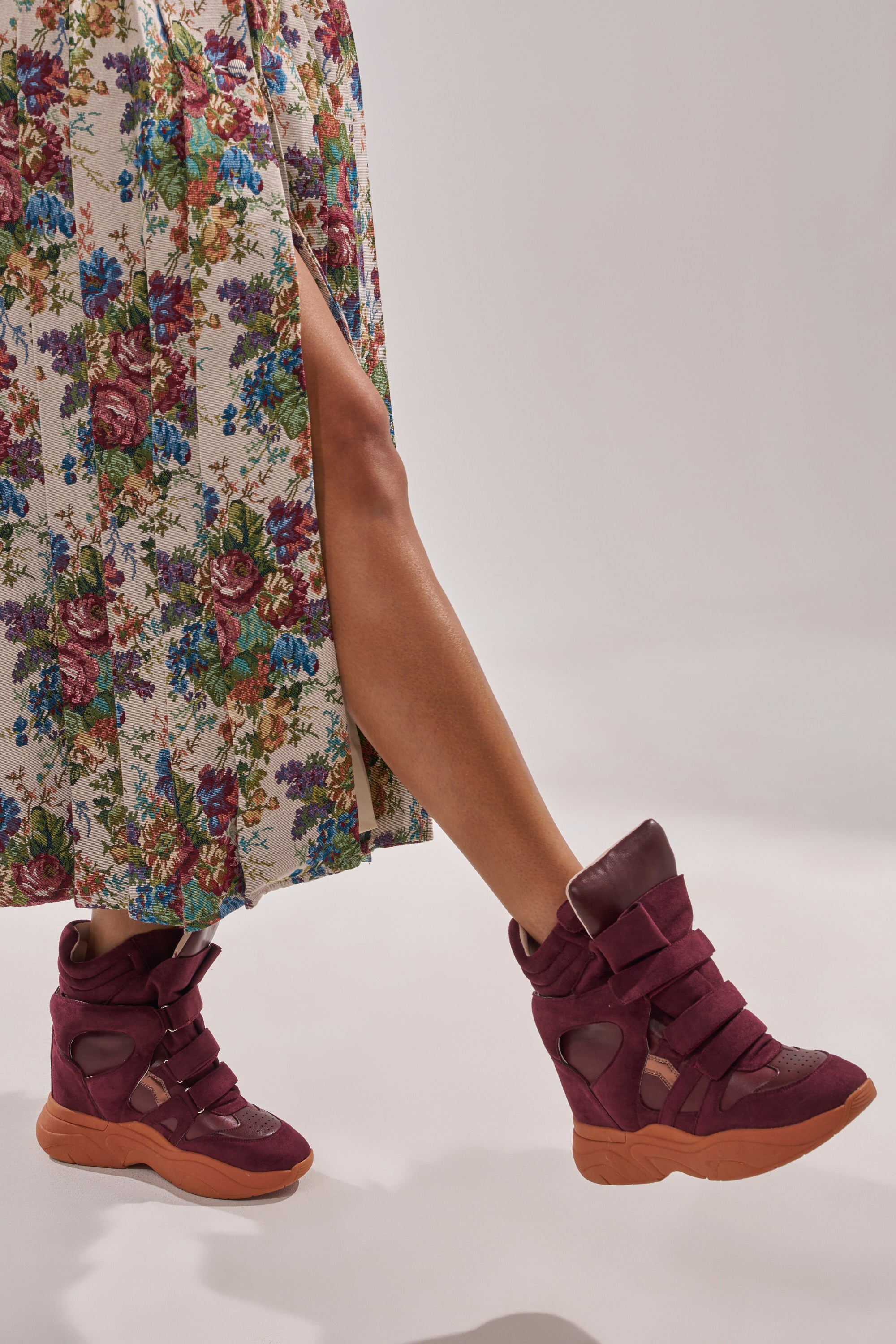 A person in a floral high-slit skirt walks, revealing one bare leg and wearing MARANTO BURGUNDY WEDGE FAUX SUEDE SNEAKER against a plain light background. 3