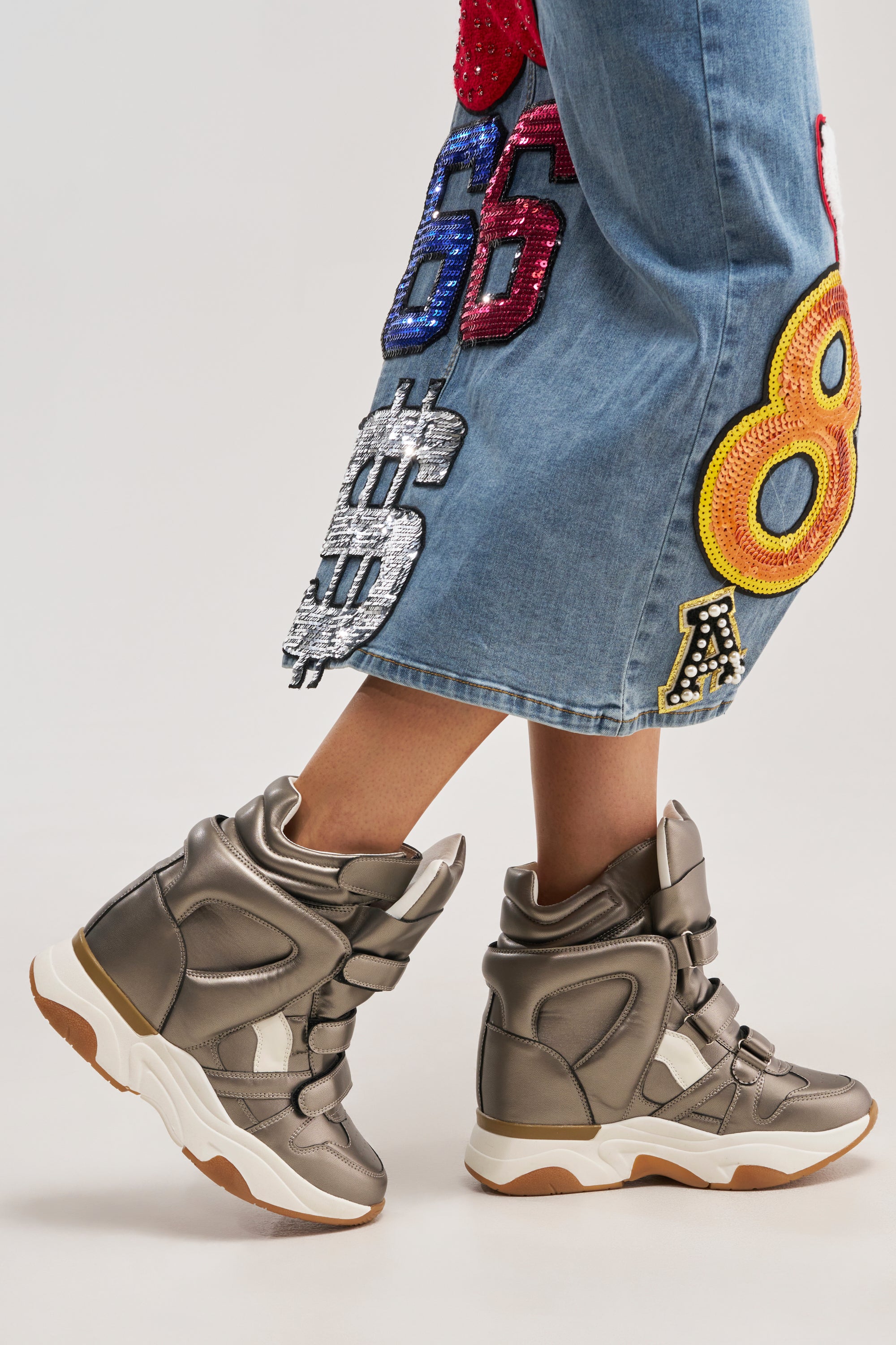 Wearing the AZALEA WANG MARANTO PEWTER WEDGE SNEAKER and blue denim pants with bold, colorful embroidered patches featuring numbers and symbols, a person walks against a plain background.