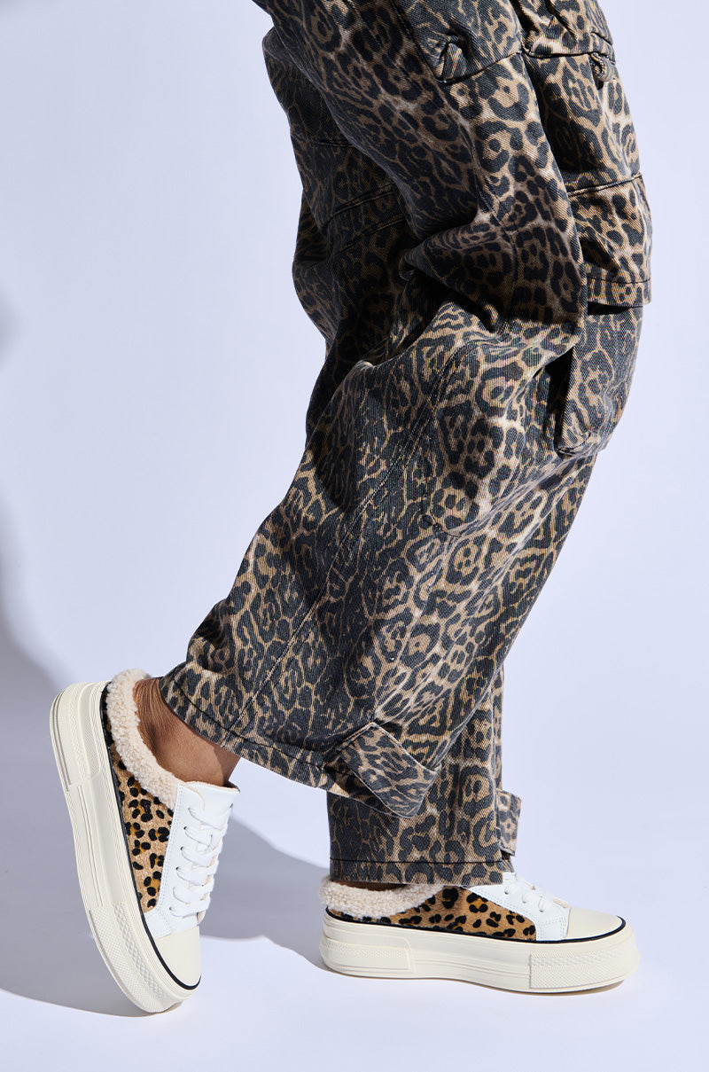 A person wearing leopard-print pants and AZALEA WANG MARGARITA FLAT SNEAKER, which has cheetah-print accents and fur trim, is walking against a light background. The image focuses on the lower legs and feet, highlighting the chic style of this sneaker.