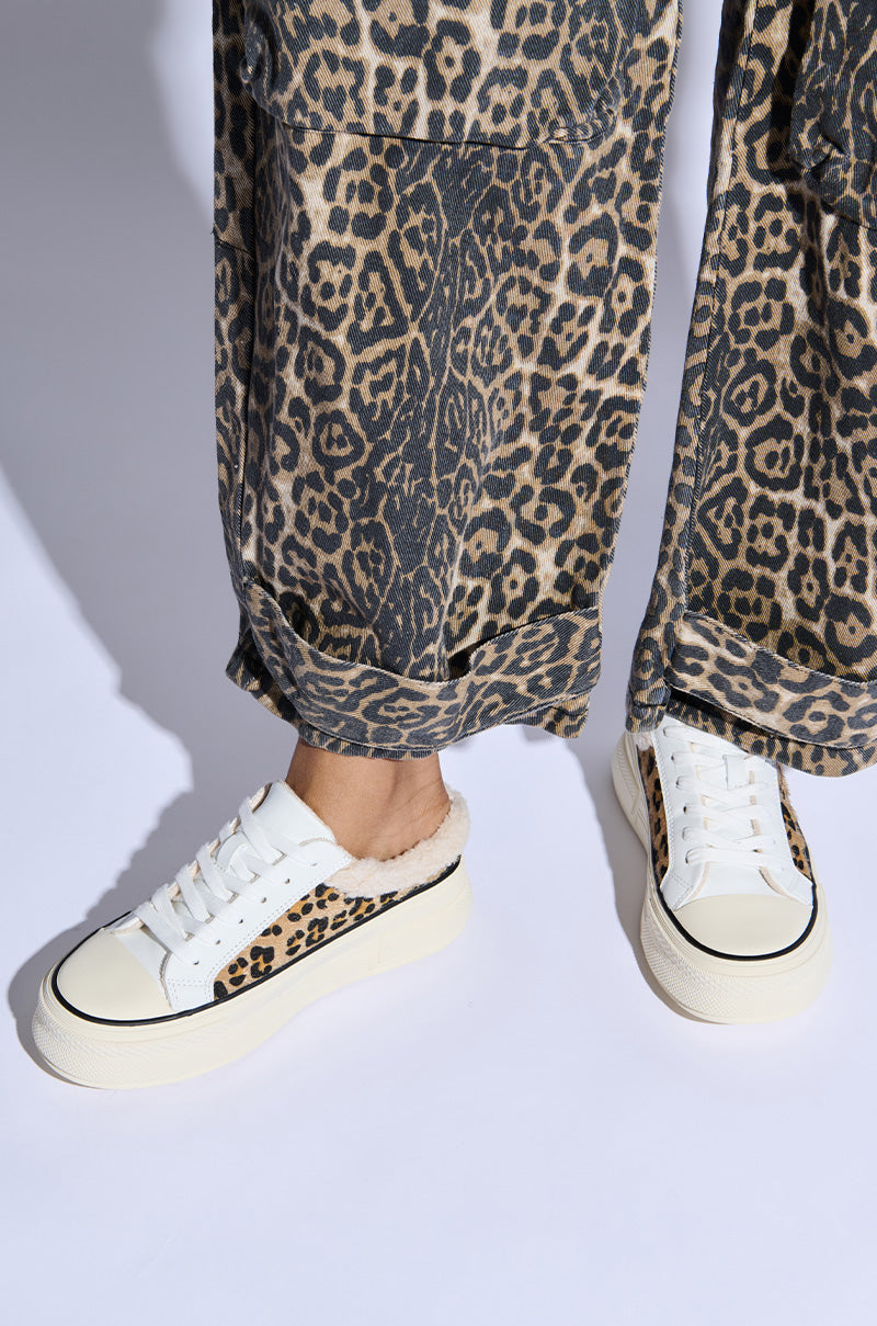 A close-up shot features a person sporting the AZALEA WANG MARGARITA FLAT SNEAKER, showcasing its white faux leather exterior with chic leopard print accents and a cozy furry lining. Paired with their leopard print pants, both feet are visible from the shins down, elegantly posed against a pristine white backdrop.