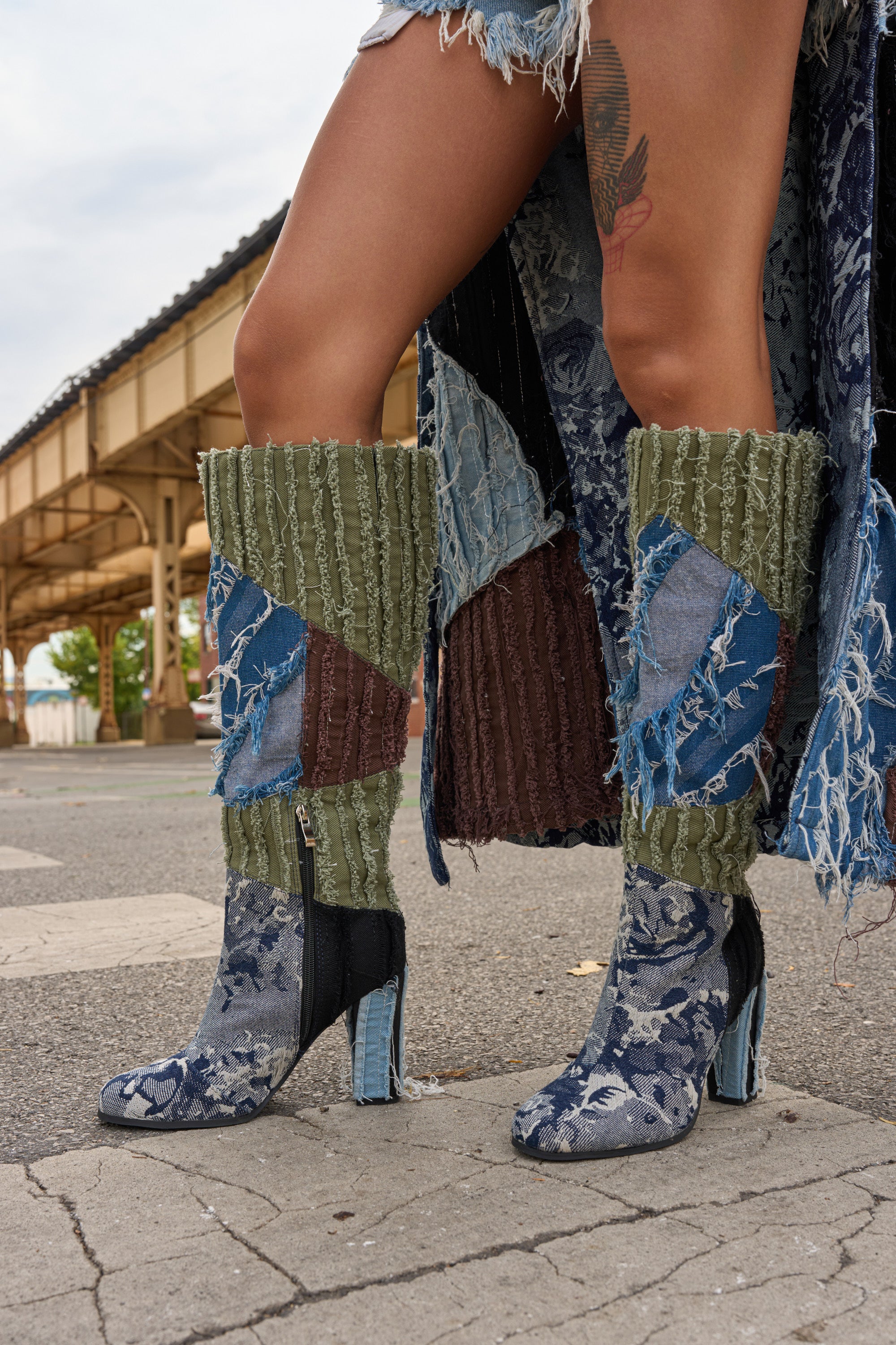 A person in the AZALEA WANG MARGEE MULTI CHUNKY KNEE HIGH BOOT, featuring patchwork green and brown fabric, stands on a street in front of train tracks. They wear a distressed, fringed skirt showing a thigh tattoo under a cloudy sky.