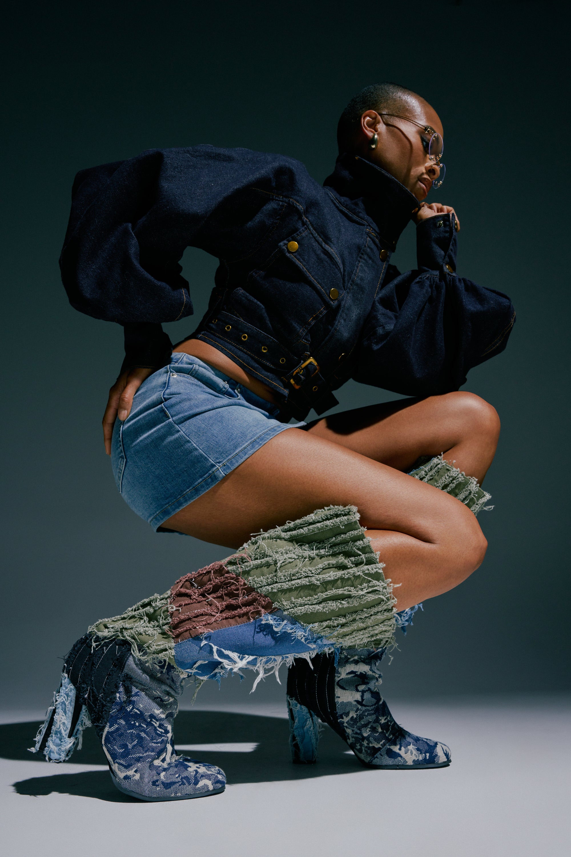 A person wearing sunglasses, a dark cropped jacket, a denim mini skirt, and the AZALEA WANG MARGEE MULTI CHUNKY KNEE HIGH BOOT poses in a crouched position against a plain background.