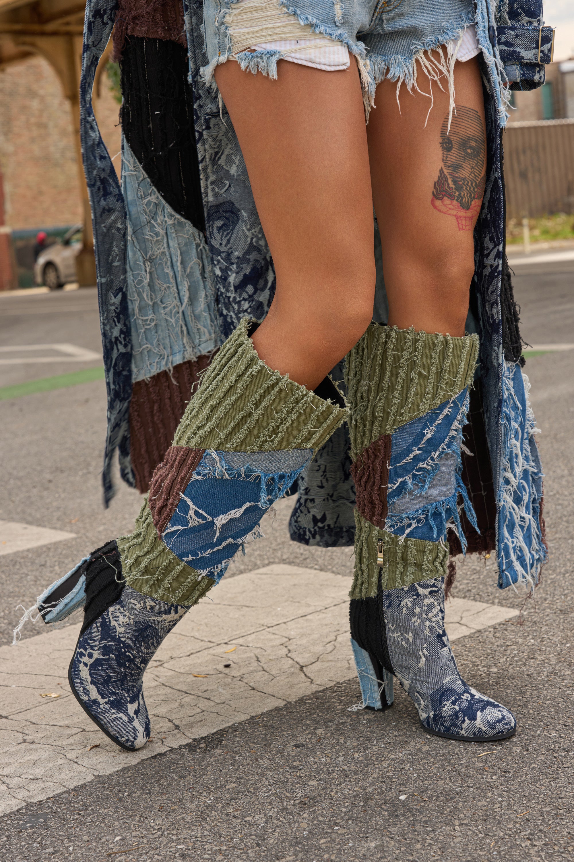 A person walks down the street in distressed denim shorts, a patchwork denim coat, and AZALEA WANG MARGEE MULTI CHUNKY KNEE HIGH BOOTS. A face tattoo on one thigh draws attention to their bold legs and standout boots.