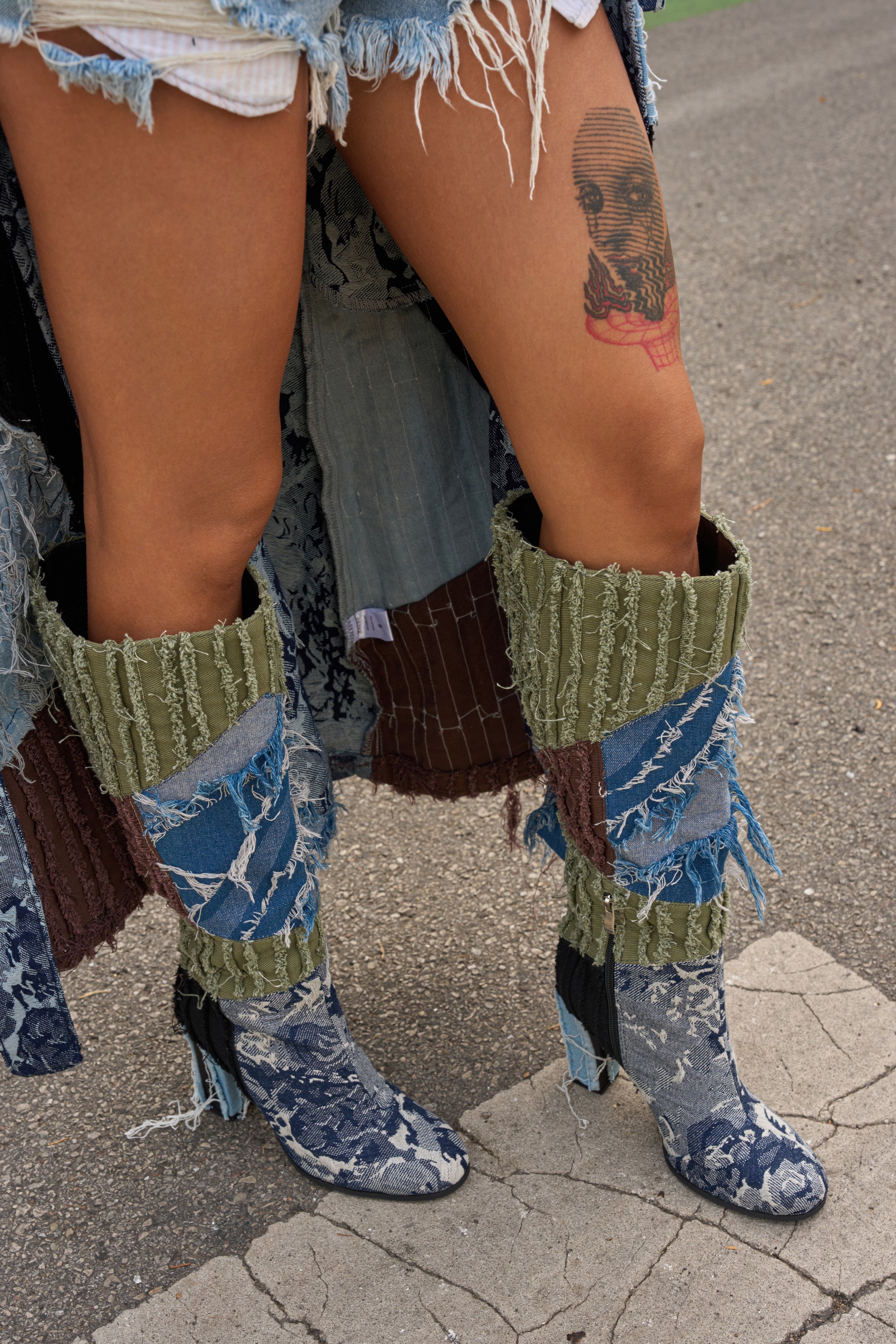 A person stands on pavement wearing distressed denim shorts and AZALEA WANG MARGEE MULTI CHUNKY KNEE HIGH BOOTS with green and blue patchwork panels, showing off a large skull thigh tattoo.