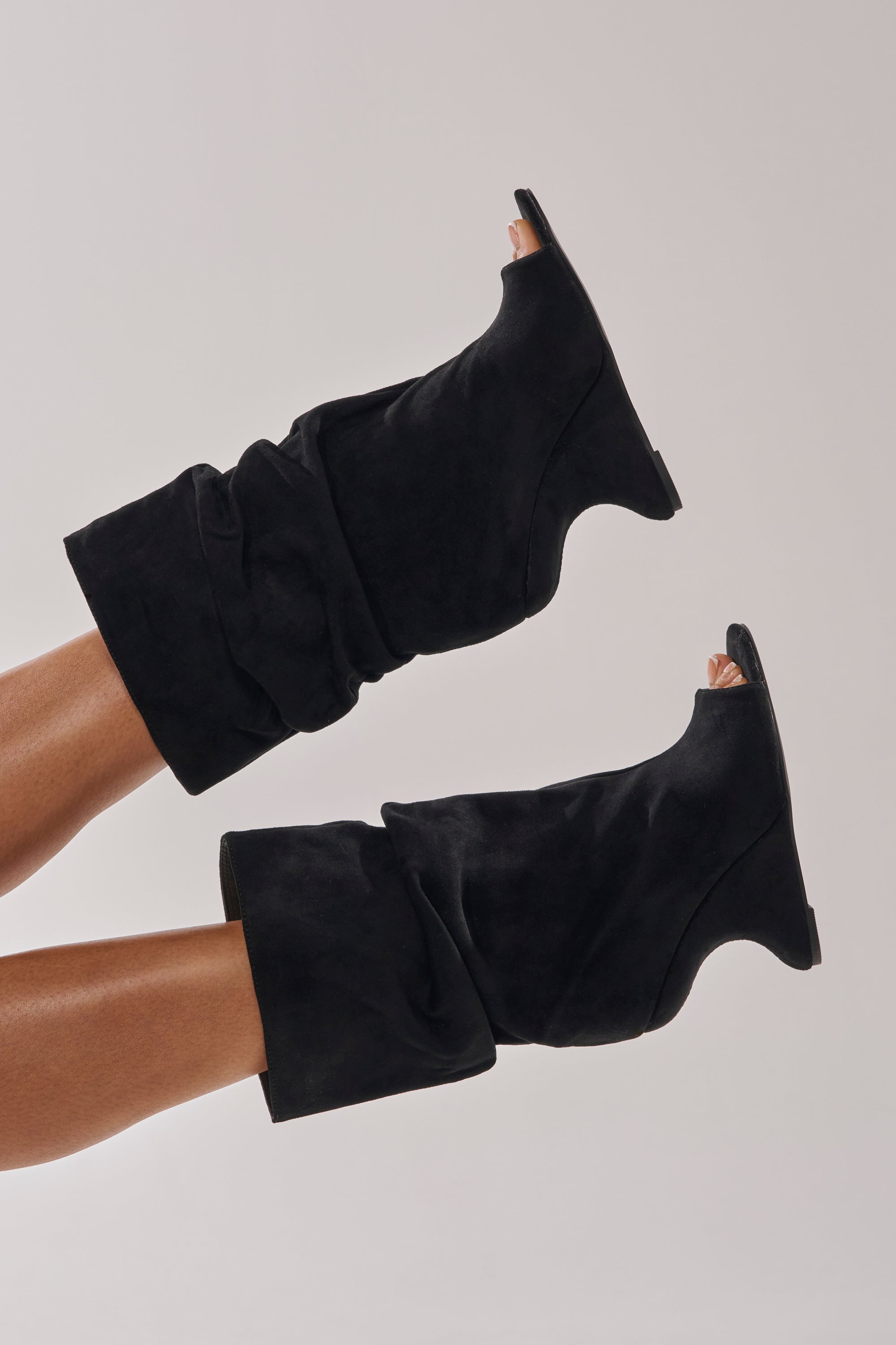 Two legs raised against a plain background, wearing the AZALEA WANG MARIN BLACK FAUX SUEDE WEDGE BOOTIE with a slouchy, relaxed fit and open-toe design. These mid-calf peep-toe booties add effortless style to any look. 0