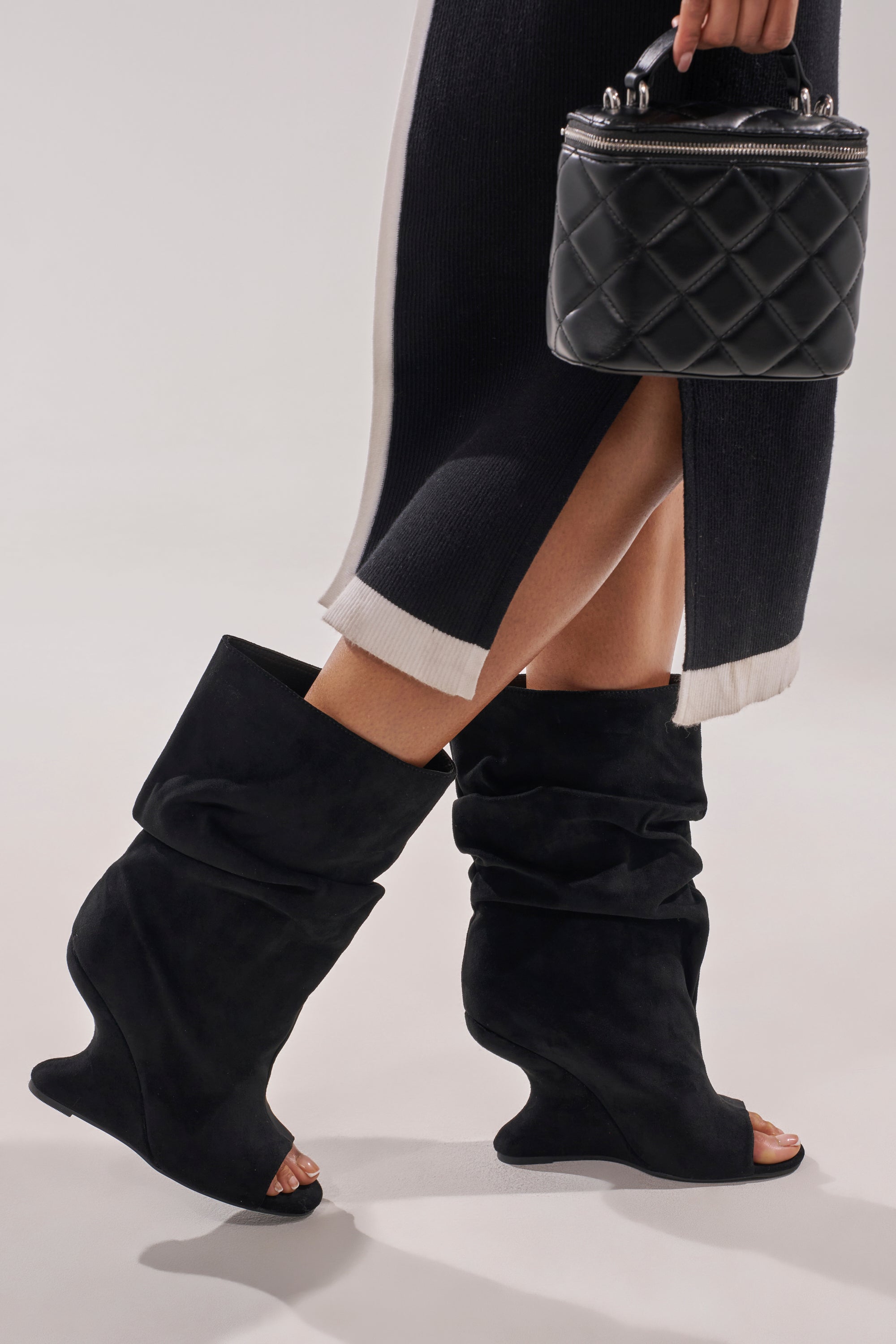 A person wearing the AZALEA WANG MARIN BLACK FAUX SUEDE WEDGE BOOTIE and a black skirt with white trim holds a quilted black top-handle handbag. 2