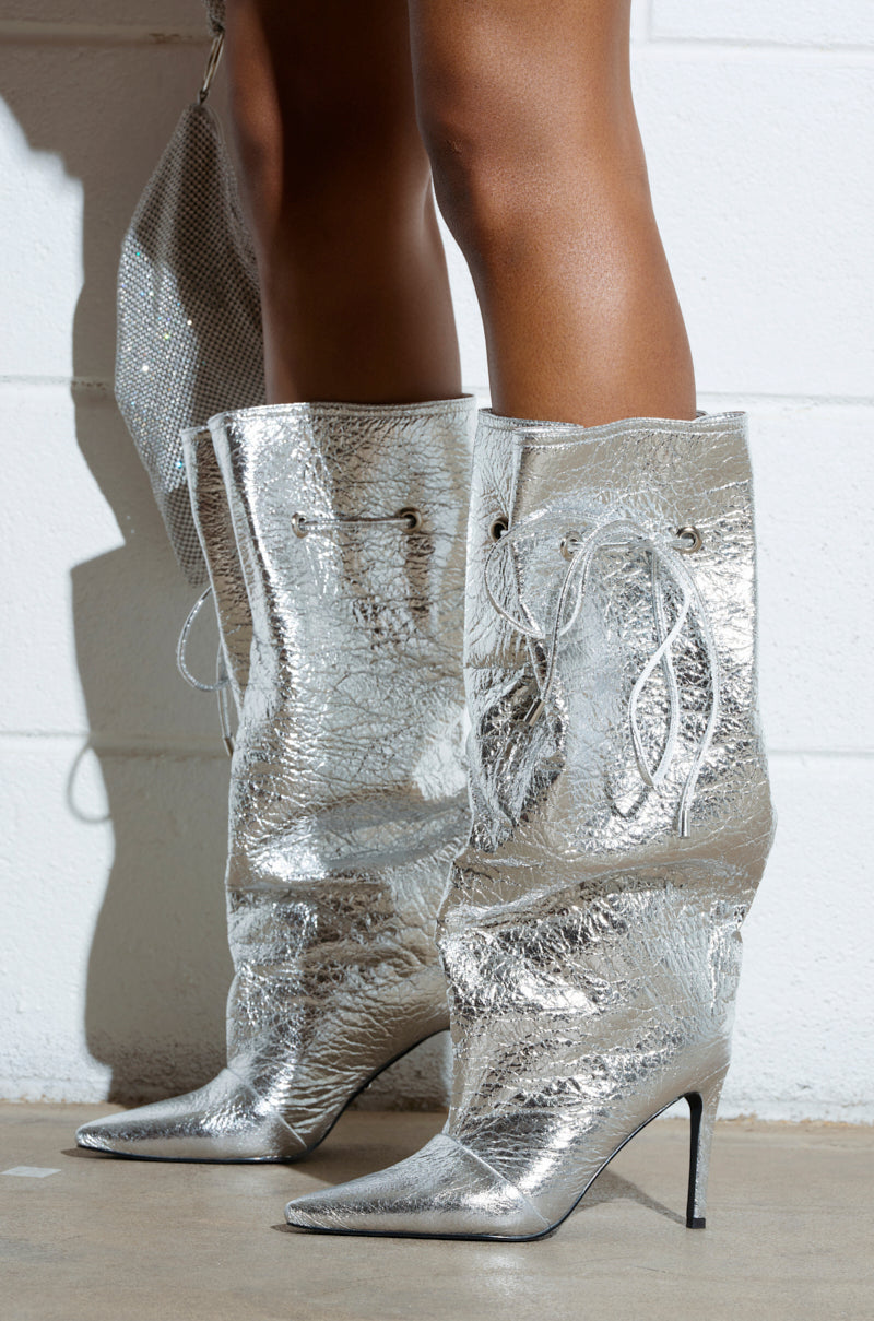 A person stands on a concrete floor wearing AZALEA WANG MARIT Silver Crackle Boots featuring a cinching mechanism and thin stiletto heels. Against the textured white wall, a matching silver bag is partially visible.