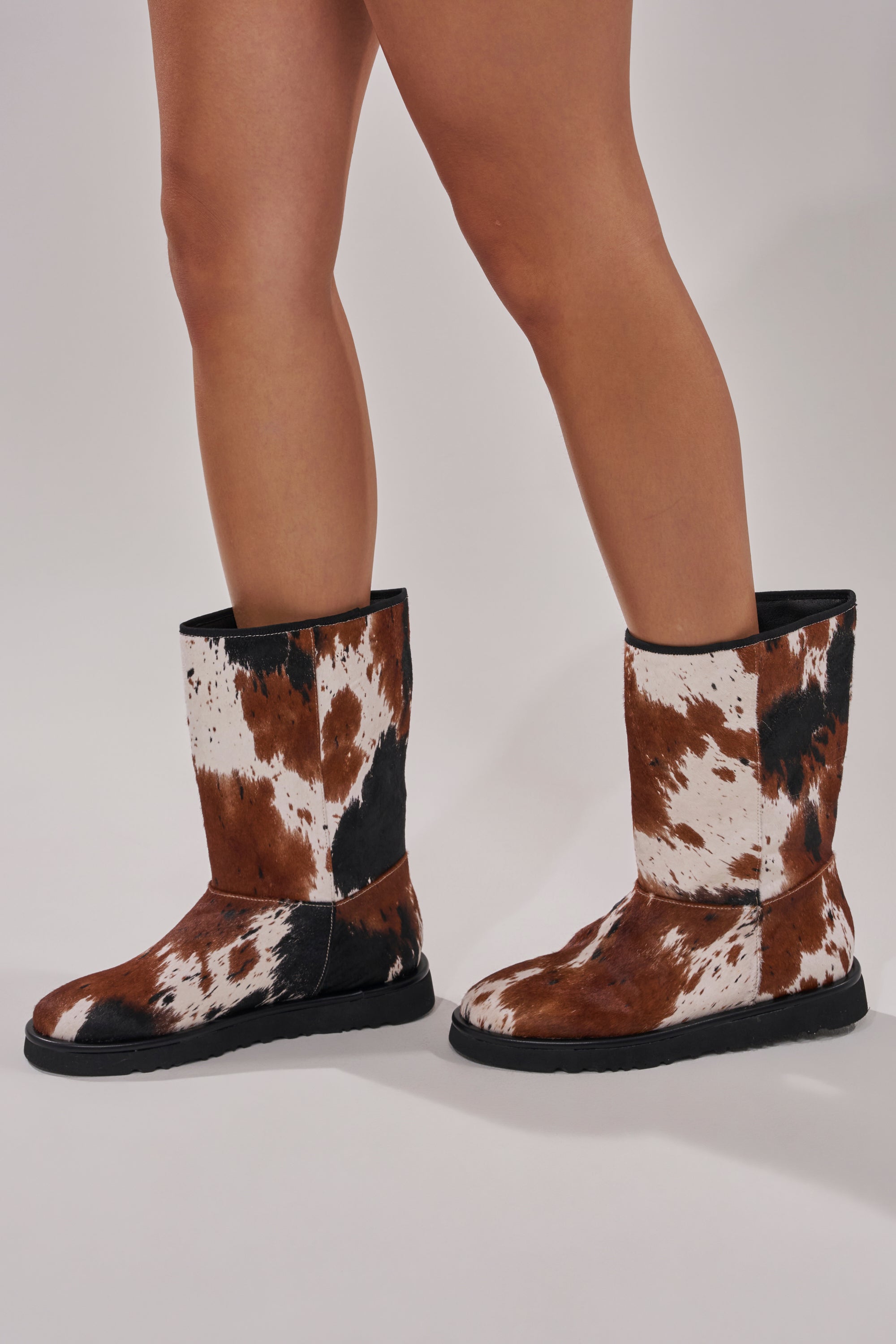 A person wears the AZALEA WANG MARLEIGH COW FLAT BOOTIE, made from genuine calf hair with brown, white, and black cow print patterns, standing on a light grey background.