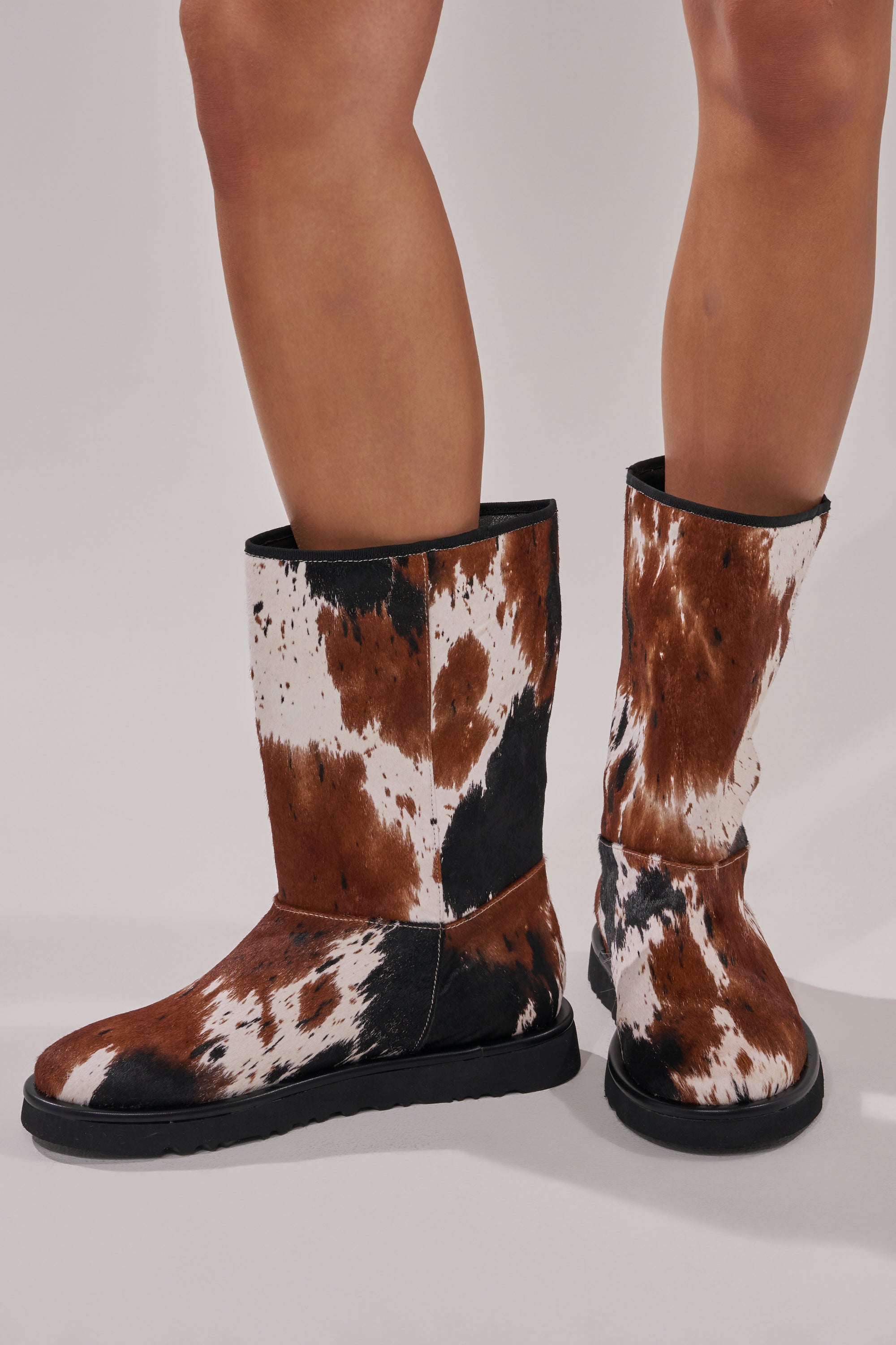 A person wearing AZALEA WANG MARLEIGH COW FLAT BOOTIE—flat mid-calf boots with a brown, black, and white cow print, black soles, and trim—stands on a light gray surface.