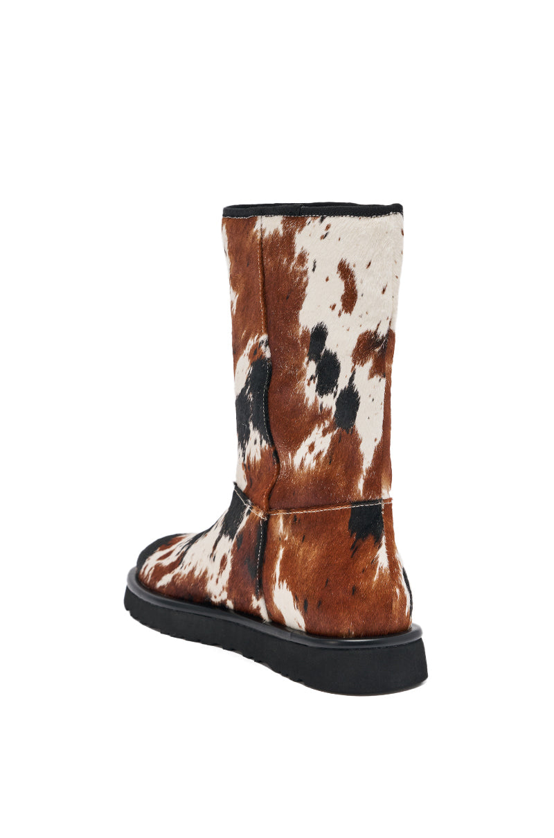 The AZALEA WANG MARLEIGH COW FLAT BOOTIE is a calf-high, flat sole bootie made from genuine calf hair with a black base and a brown, black, and white cow print, shown from a rear side angle on a white background. 5