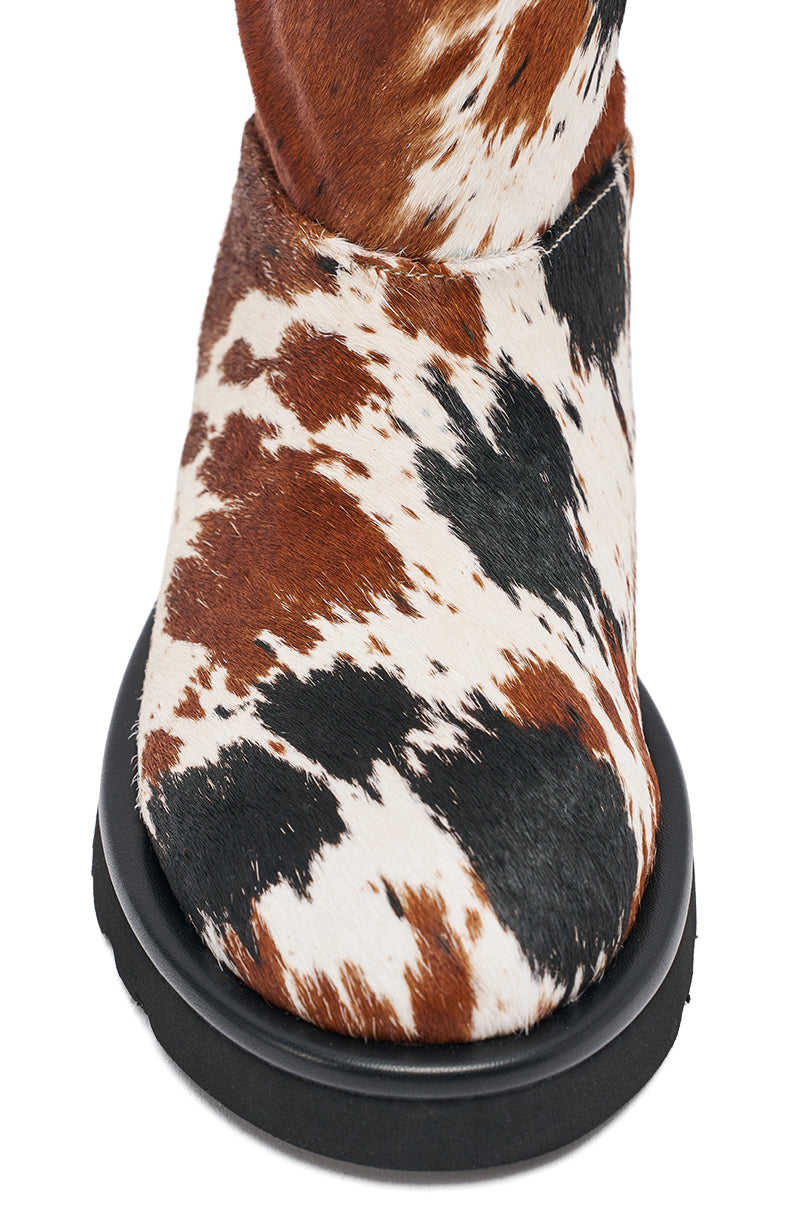 A close-up of the AZALEA WANG MARLEIGH COW FLAT BOOTIE, featuring genuine calf hair in a brown, black, and white cow print with a thick black sole. 6
