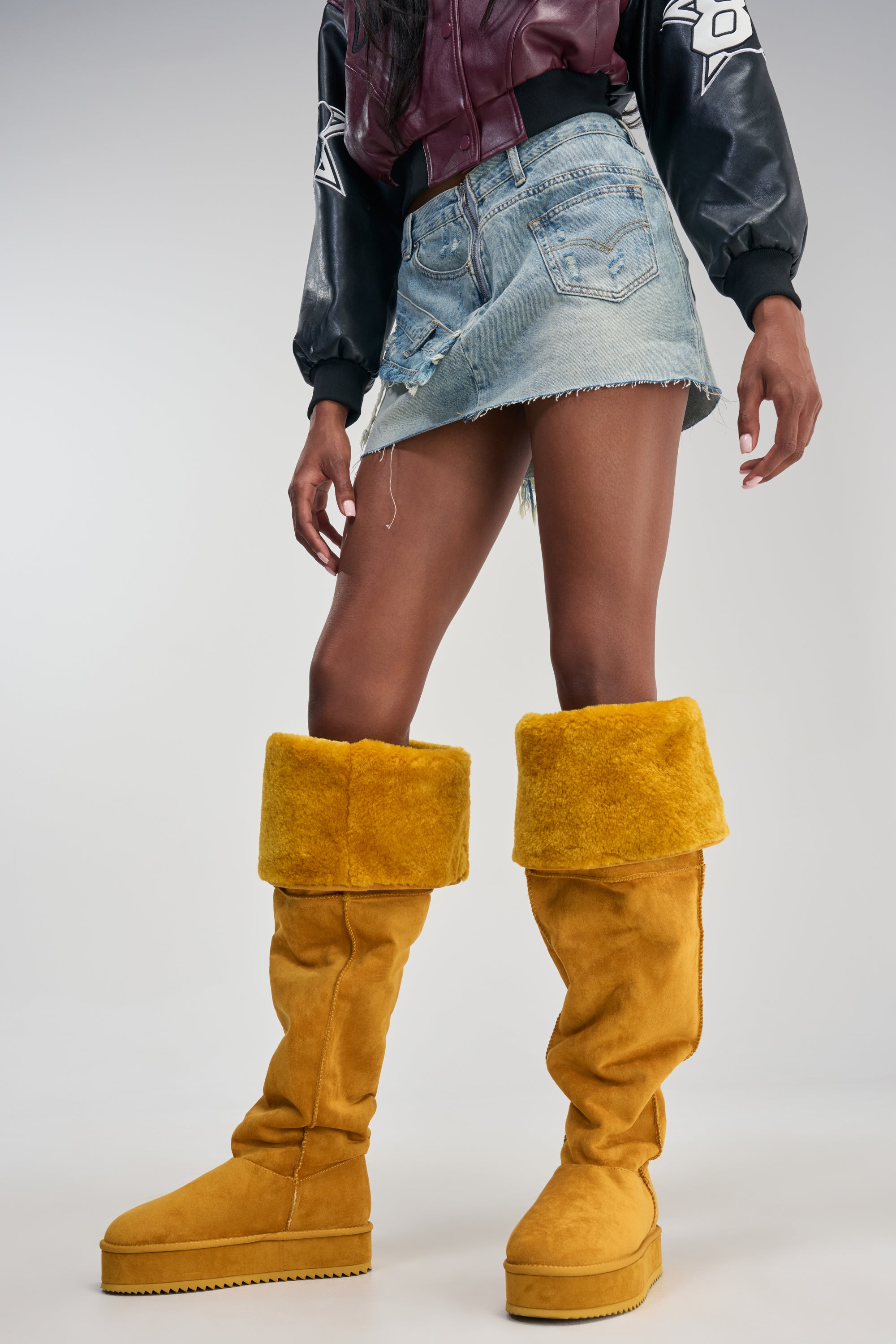 A person wearing a cropped leather jacket, a denim mini skirt, and the AZALEA WANG MARLOW MUSTARD SUEDE SHERPA FLATFORM BOOT stands against a light background.