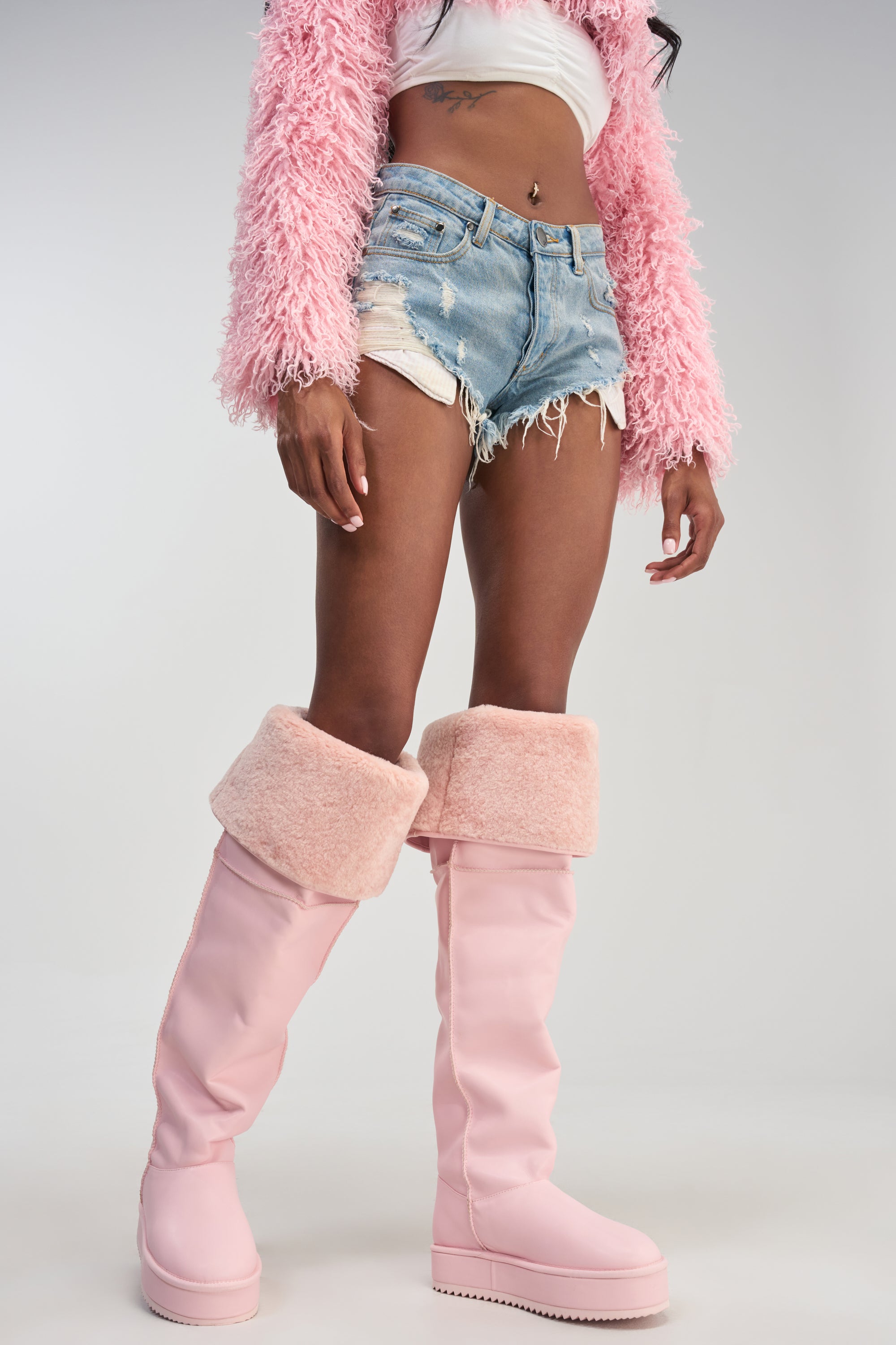 A person stands against a plain background wearing a pink fuzzy jacket, white crop top, ripped denim shorts, and the AZALEA WANG MARLOW Pink PU Sherpa Flatform Boots—knee-high pink faux leather boots with furry cuffs.