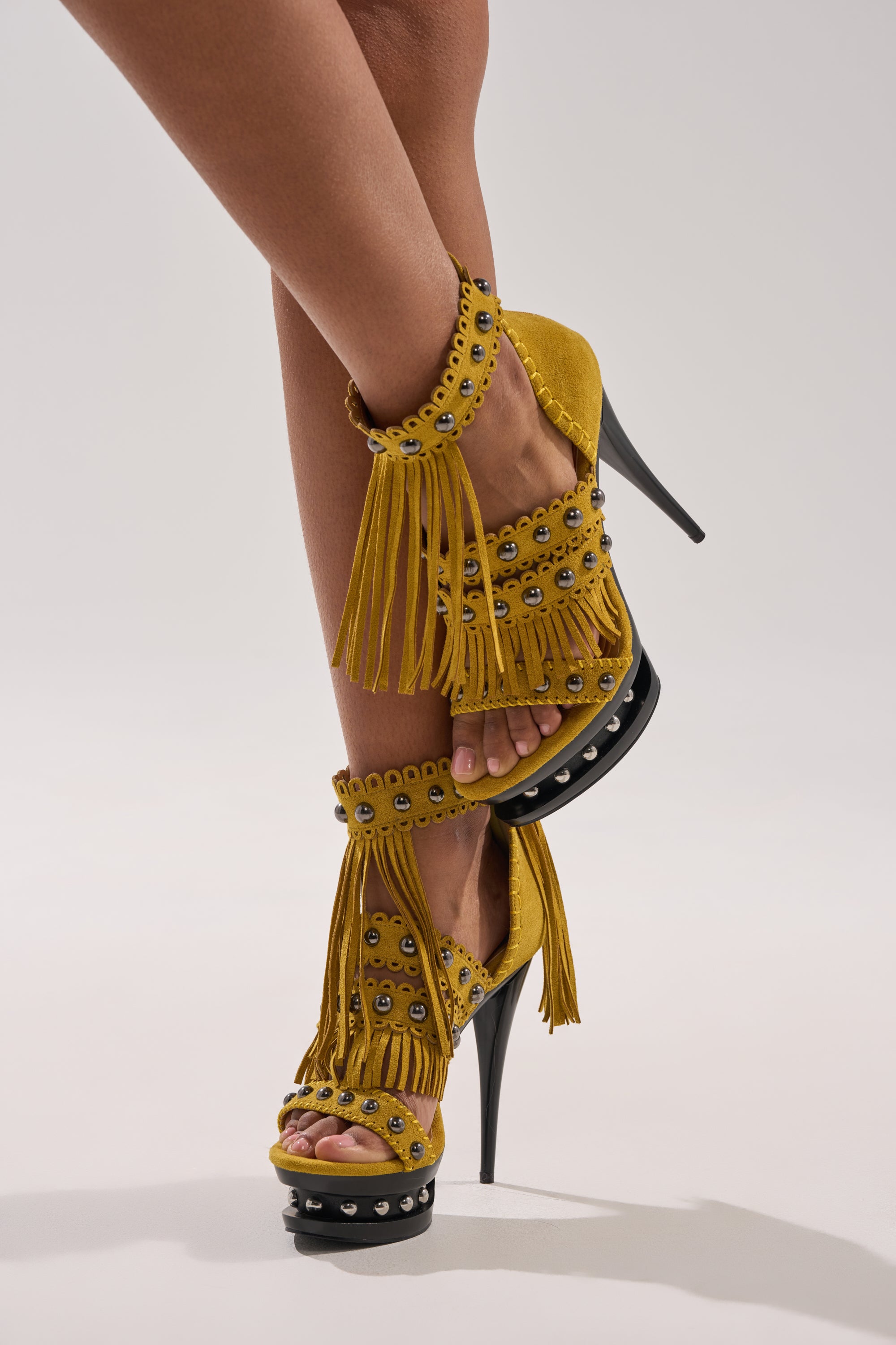 A person models the AKIRA GOLD MAROVA MUSTARD NOVELTY PLATFORM FRINGE EMBELLISHED SANDAL, featuring metallic studs and bold mustard fringe, while crossing one leg over the other against a plain light background. 0