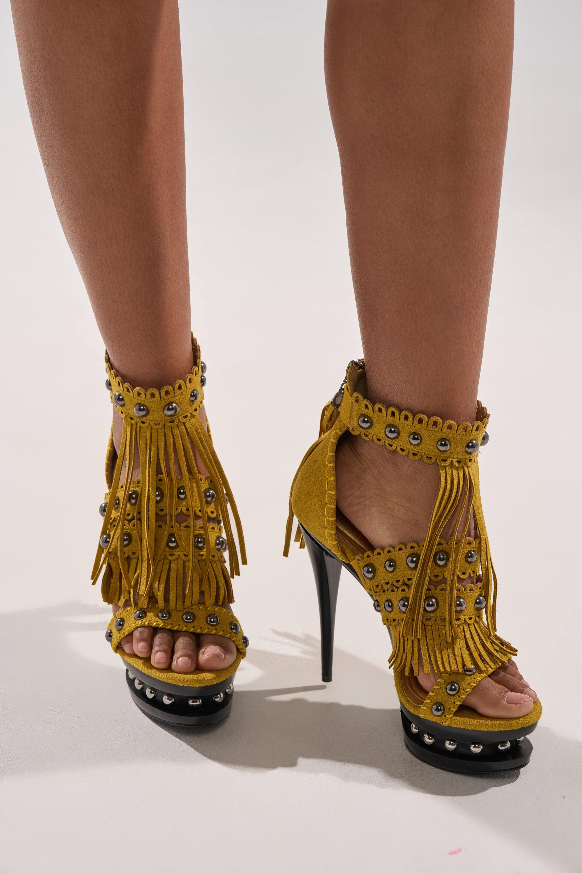 A close-up of a person wearing the AKIRA GOLD MAROVA Mustard Novelty Platform Sandal, featuring fringe embellishments, metal eyelets, and an ankle strap, standing on a light-colored surface. 2