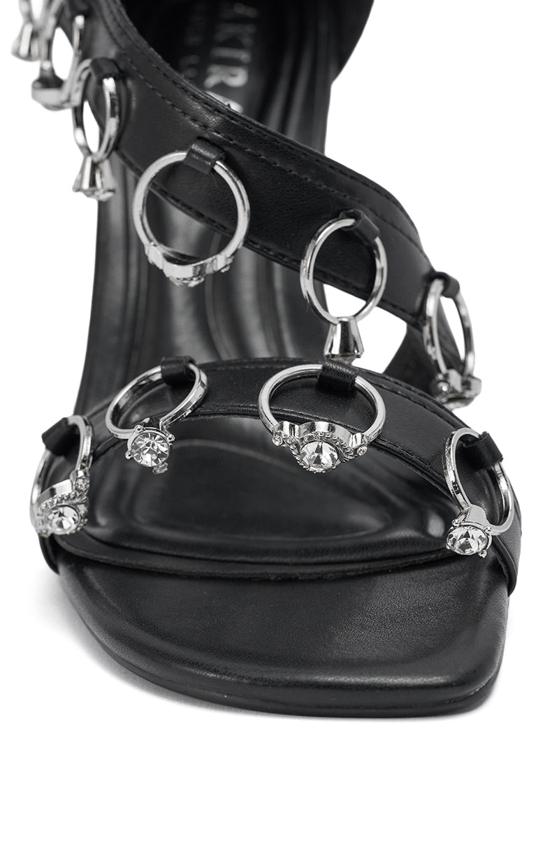Front view of the AKIRA GOLD MARQUISE BLACK STILETTO SANDAL WITH RINGS, featuring a black faux leather design, open square toe, multiple silver rings, and sparkling rhinestone embellishments on the straps. 6