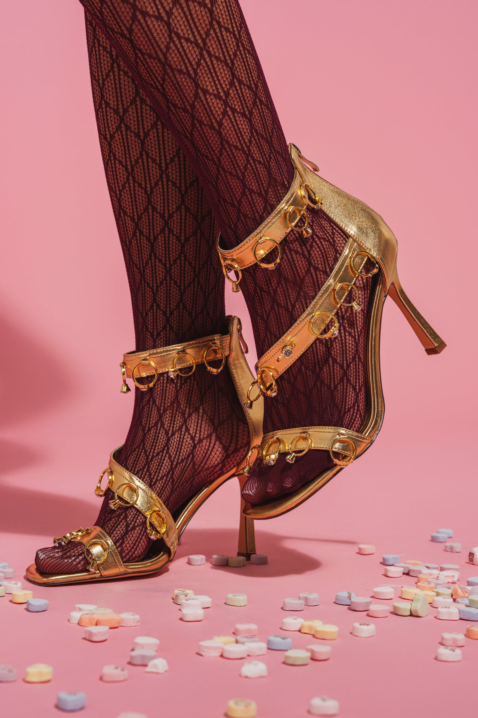 Wearing AKIRA GOLD MARQUISE GOLD STILETTO SANDAL WITH RINGS and patterned burgundy tights, a person stands on a pink surface covered with colorful candy hearts. 1