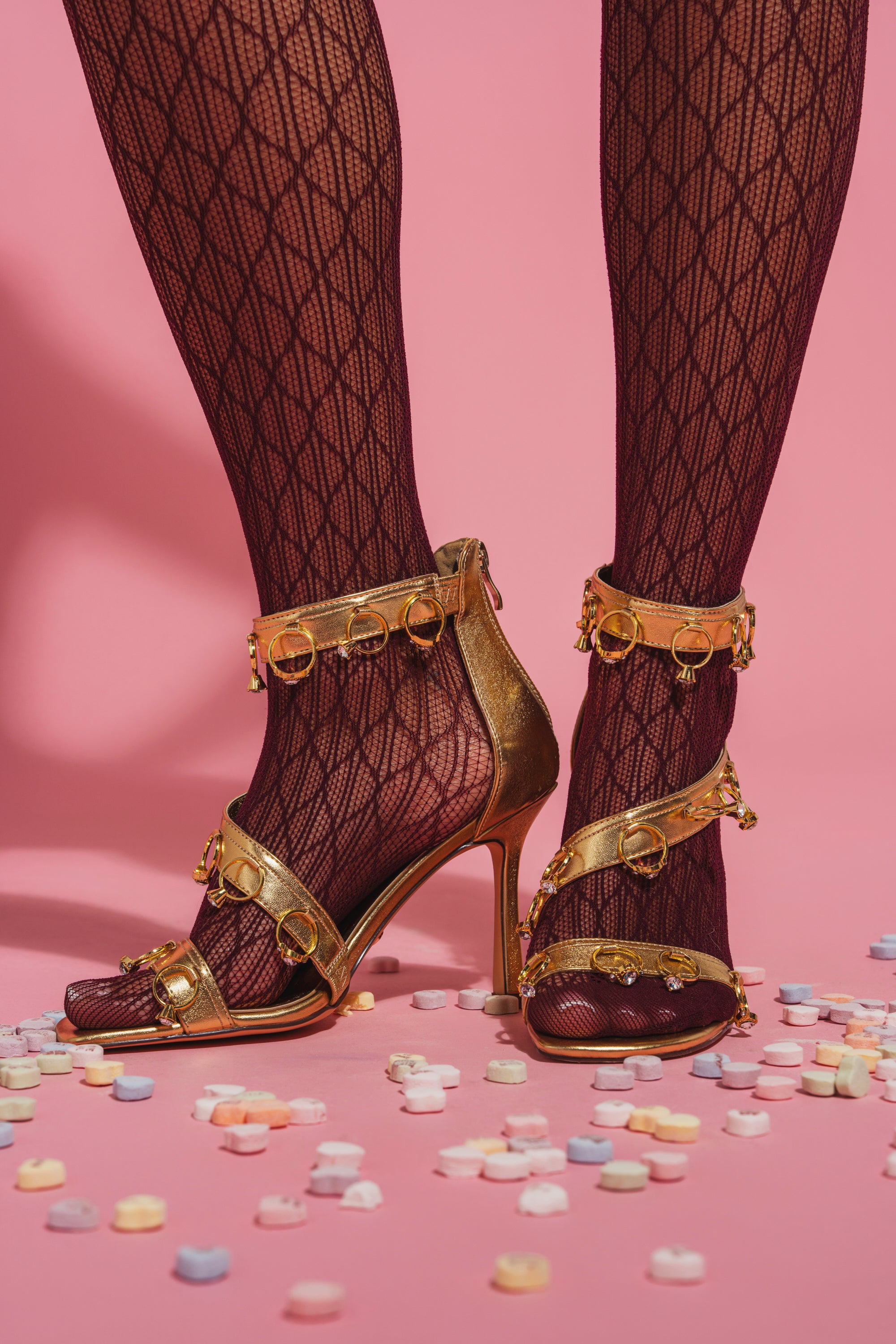 A close-up of legs in black patterned tights and the AKIRA GOLD MARQUISE GOLD STILETTO SANDAL WITH RINGS, standing on a pink surface with colorful candy hearts—an eye-catching setup to highlight bold statement heels. 2