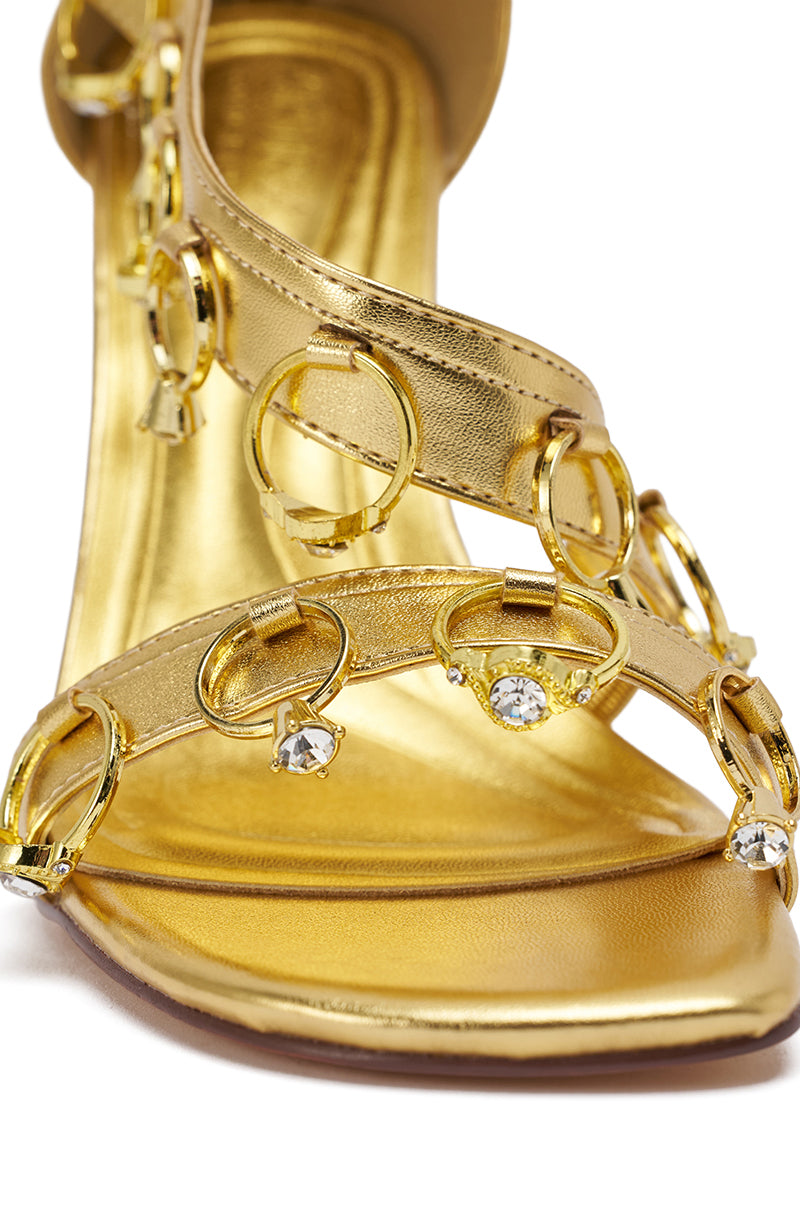 The AKIRA GOLD MARQUISE GOLD STILETTO SANDAL WITH RINGS features metallic faux leather, three straps with shiny circular rings and clear gemstones, and an elegant heel, highlighted from the front for its statement design. 9