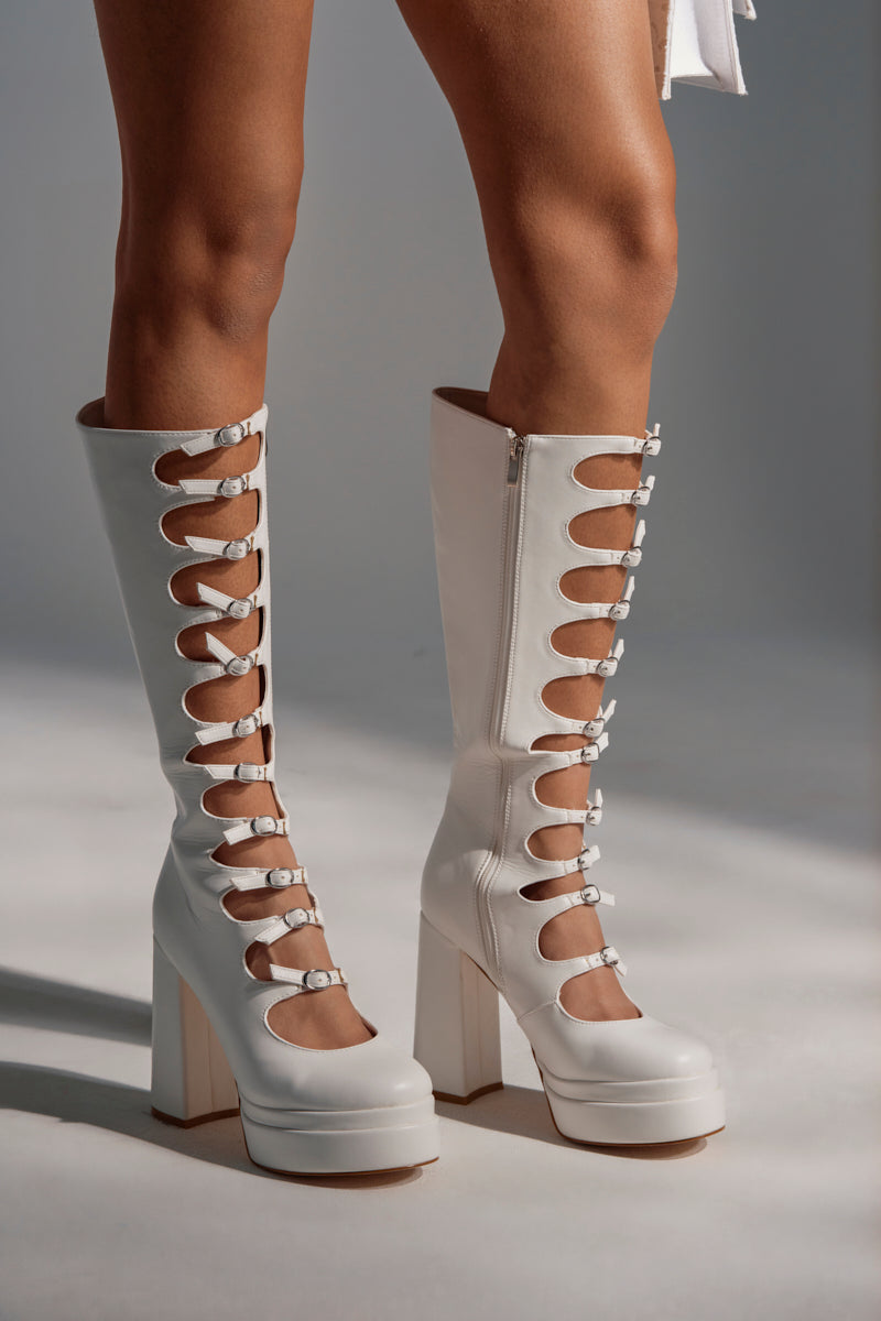 A person wears the AZALEA WANG MARRIGOLD BOOT IN WHITE, featuring knee-high, white faux leather with multiple circular cutouts and buckle straps, while standing on a light-colored surface.