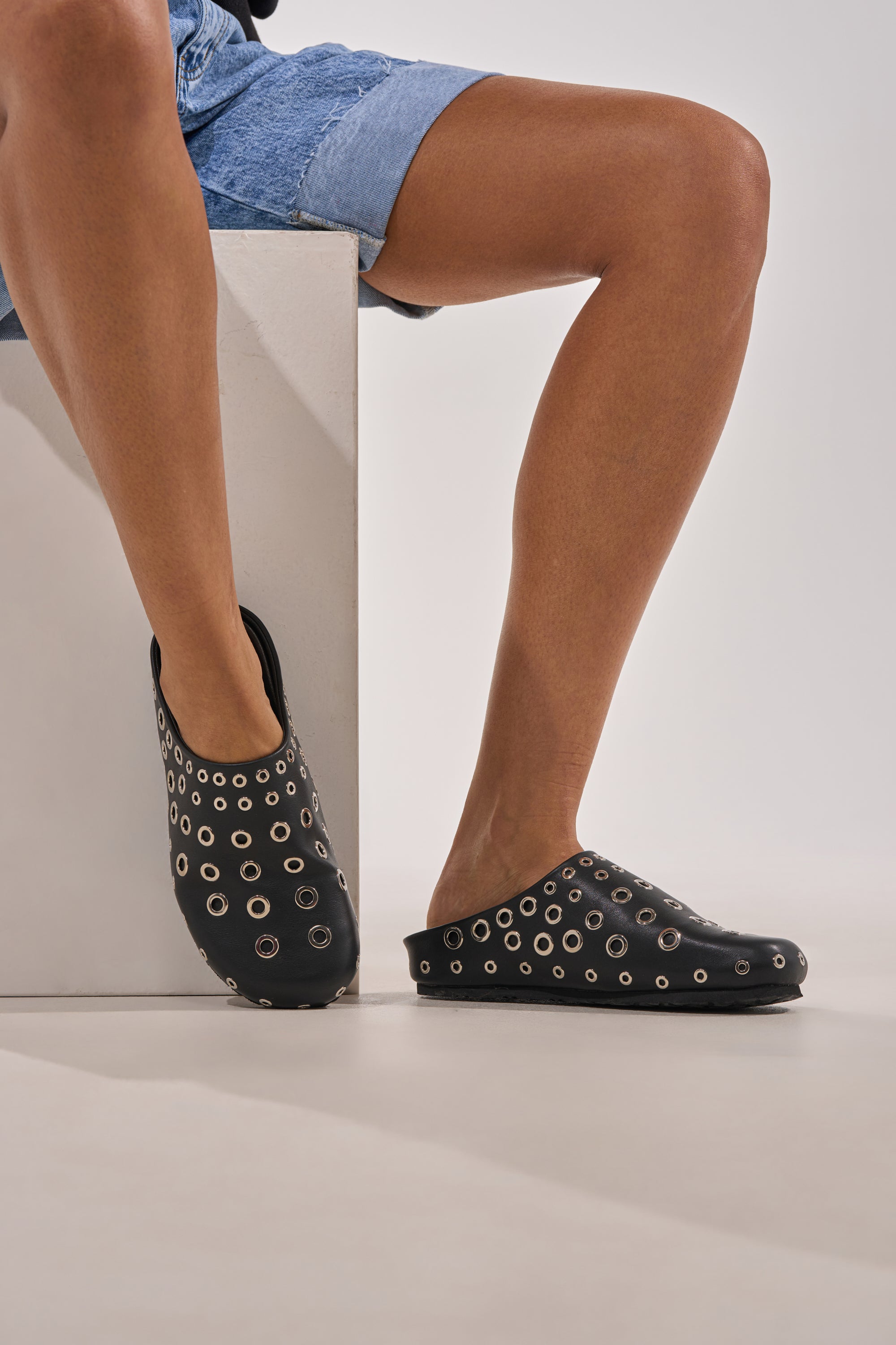 A person in denim shorts crosses their legs, showcasing the AZALEA WANG MARSHA BLACK GROMMET DETAIL FLAT—sleek black slip-on clogs with silver grommets that stand out stylishly against a neutral background. 0