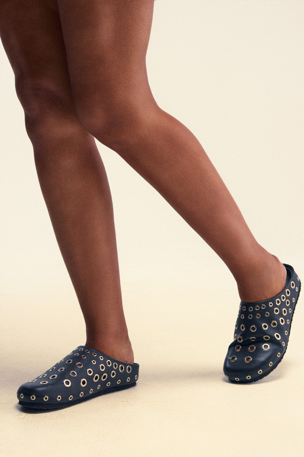 Wearing the AZALEA WANG MARSHA BLACK GROMMET DETAIL FLAT, a stylish pair of slip-on flats adorned with multiple gold grommets and reminiscent of chic faux leather clogs, a person stands with legs crossed against a neutral background.