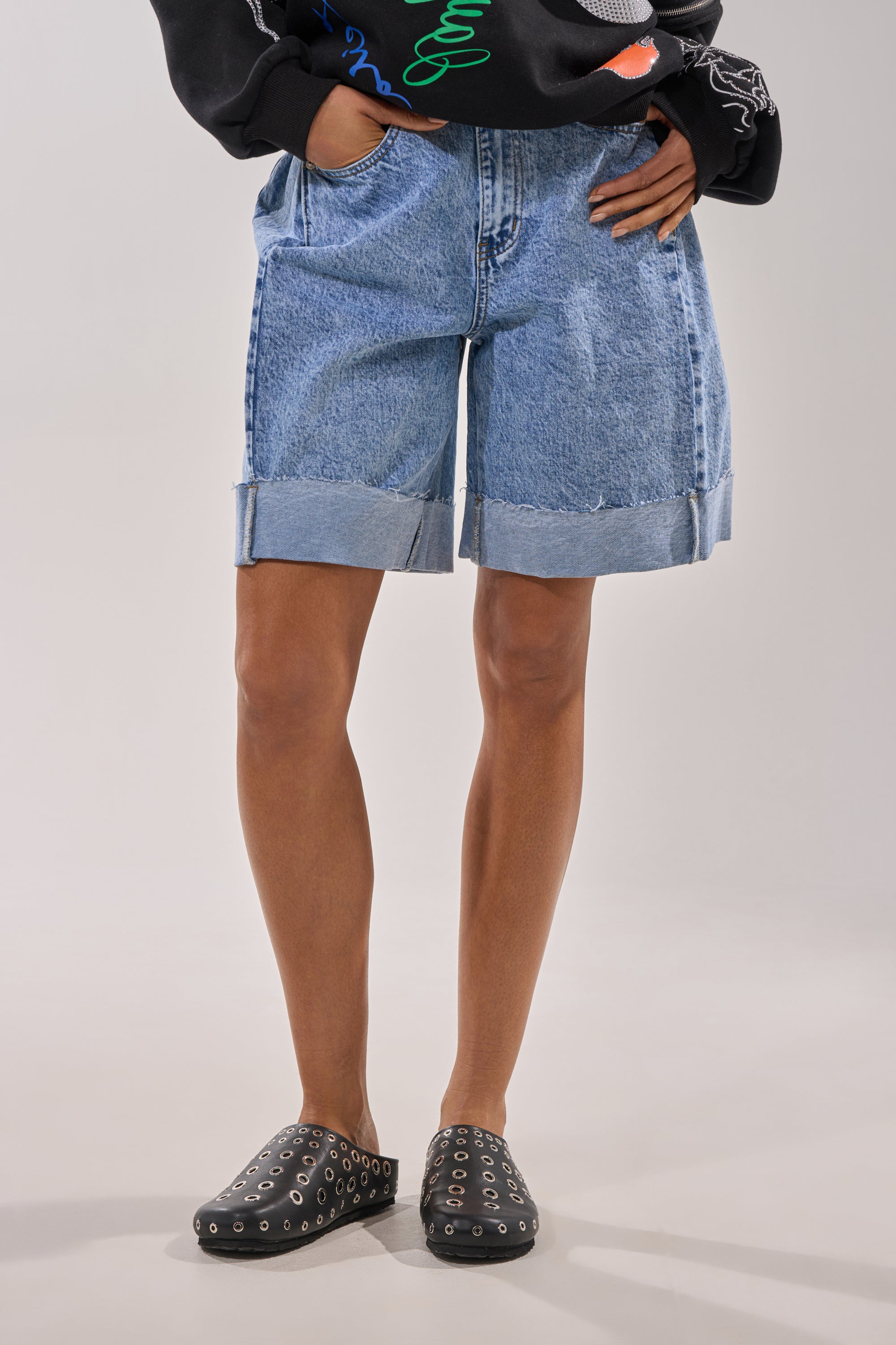 A person stands with hands in pockets, wearing a black sweater, light blue rolled-up denim shorts, and AZALEA WANG MARSHA BLACK GROMMET DETAIL FLATS. The photo is cropped at the shoulders. 1