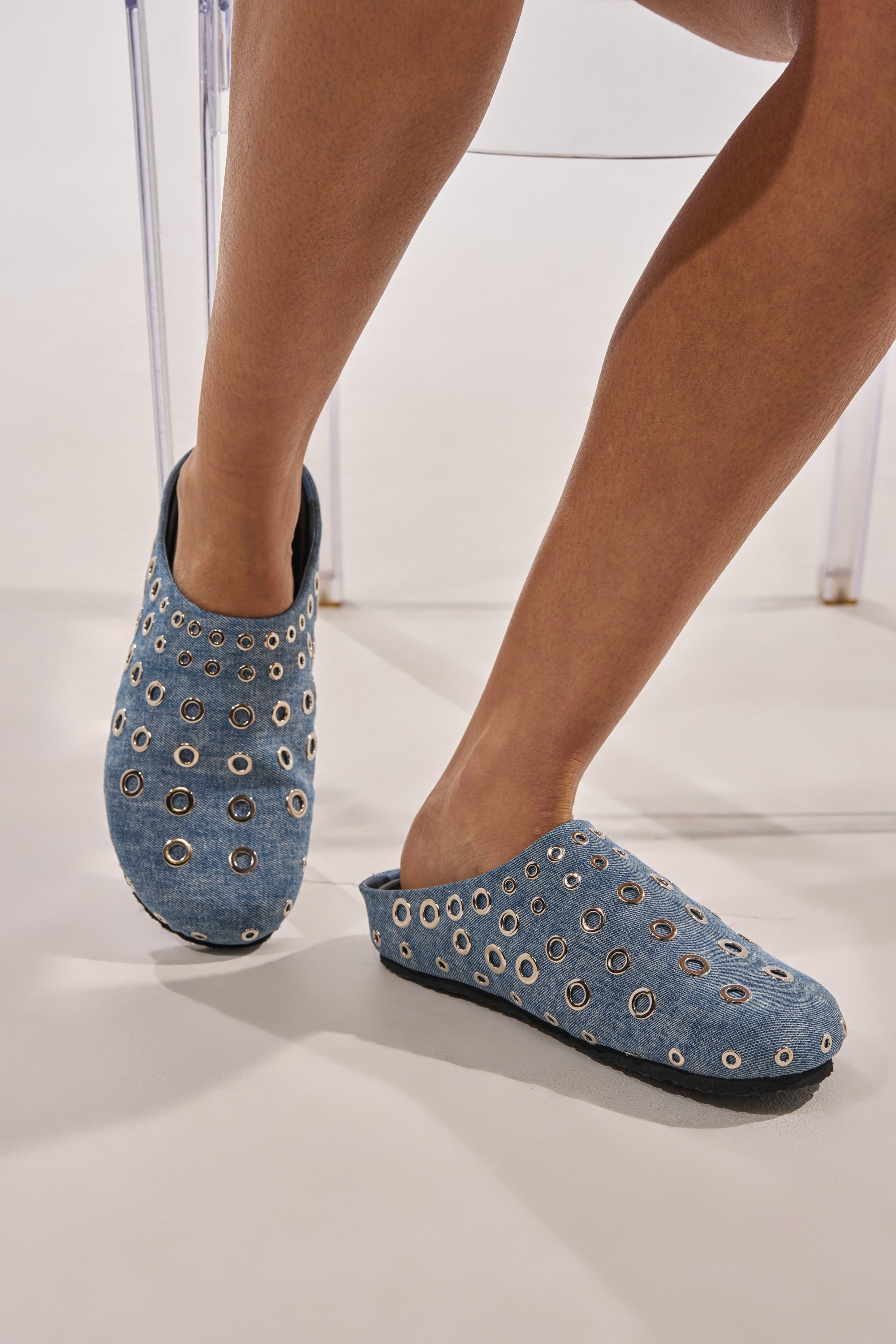 A person sits with legs crossed, wearing the AZALEA WANG MARSHA DENIM GROMMET DETAIL FLAT shoes in blue, featuring silver grommet accents, set against a neutral background. 0
