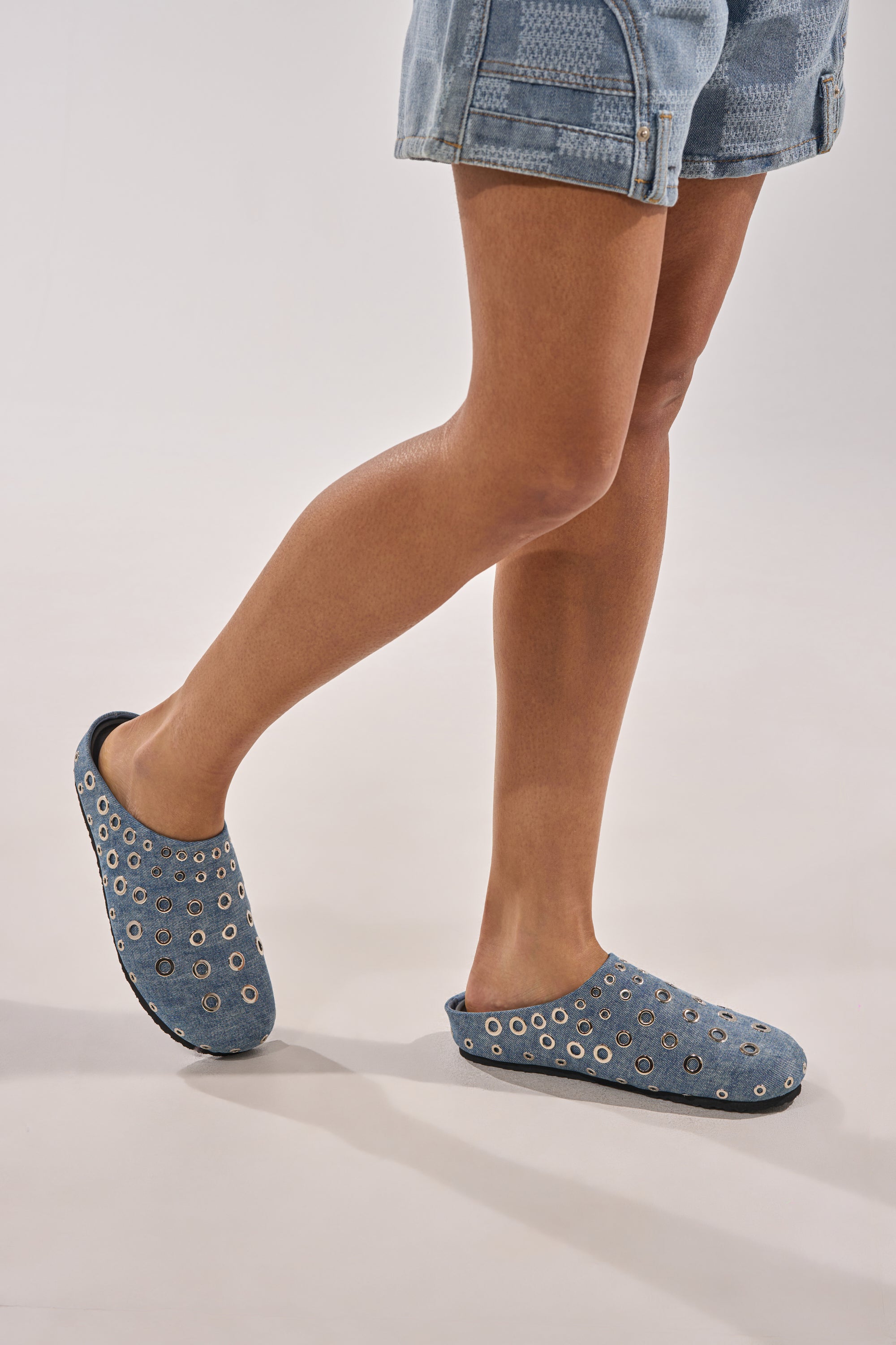 A person wearing AZALEA WANG MARSHA DENIM GROMMET DETAIL FLATS stands with one leg slightly bent, dressed in blue denim shorts against a plain light background. 1