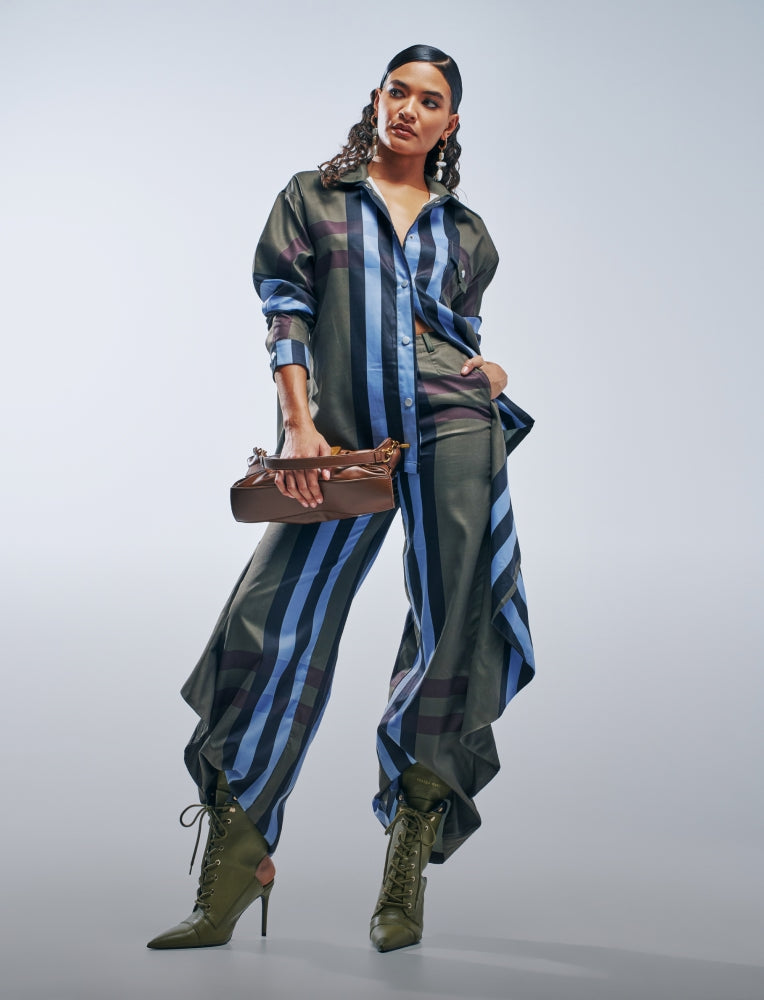 A model poses in a matching two-piece satin-like jumpsuit set featuring vertical blue, gray, and purple stripes, styled with olive green lace-up ankle boots and a brown leather clutch bag against a light gray studio background.