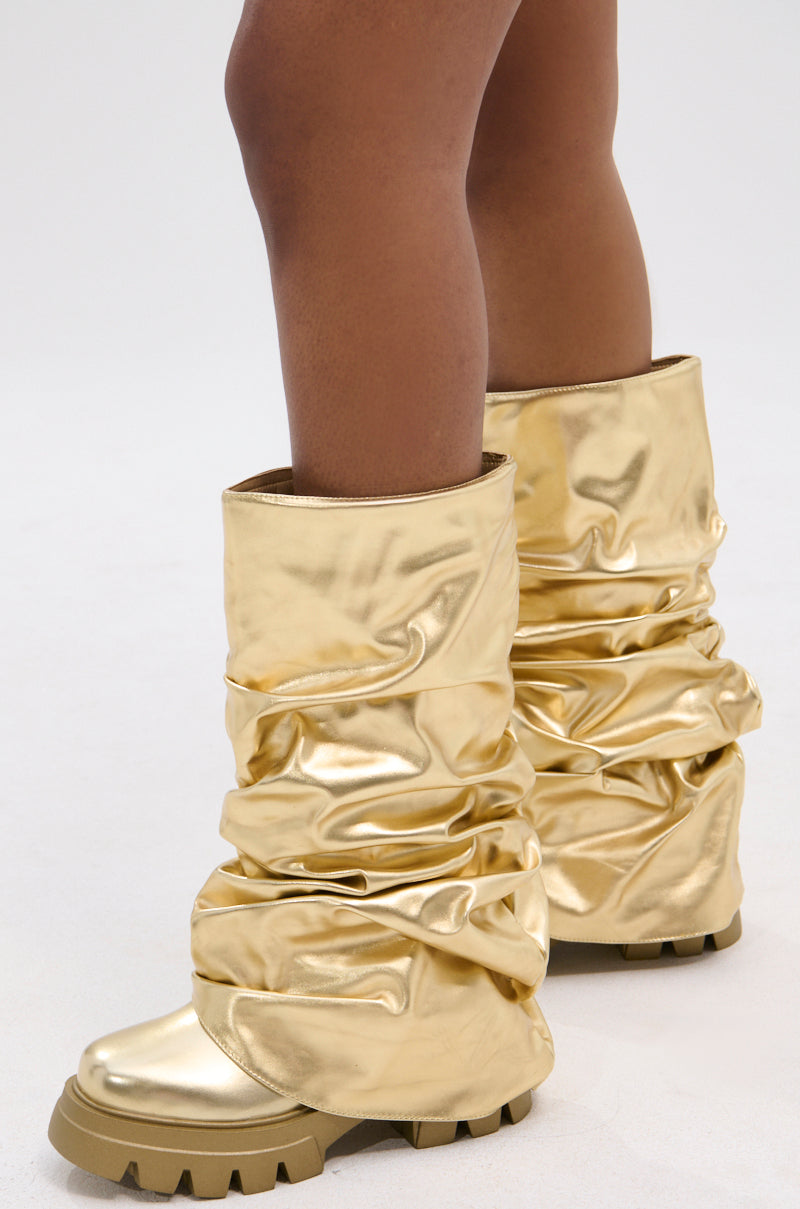 A person wears the AZALEA WANG MATE METALLIC MID CALF BOOT IN GOLD—tall, shiny gold boots with thick, rugged soles that reach the lower calf. The textured metallic finish stands out against a plain, light backdrop.