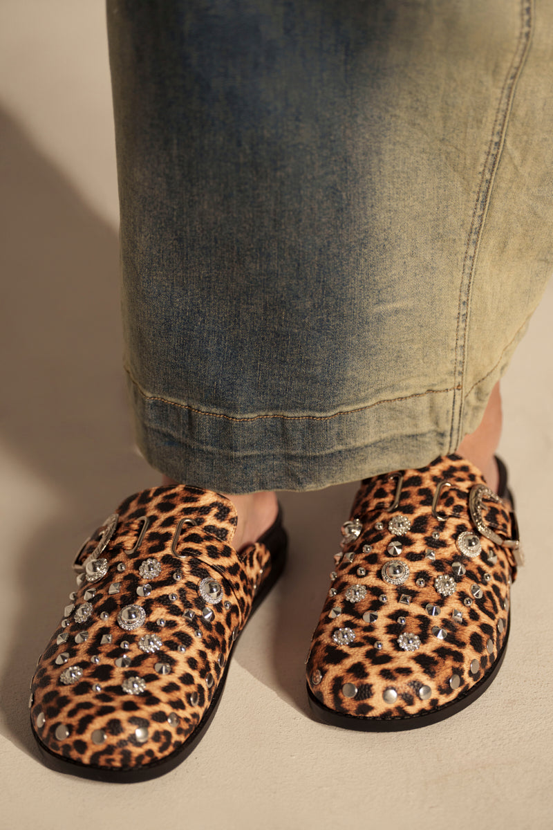 A person wearing a long, faded denim skirt and AZALEA WANG MAUDLIN LEOPARD CLOGS with faux leather accents and large, shiny rhinestones stands on a light-colored floor.