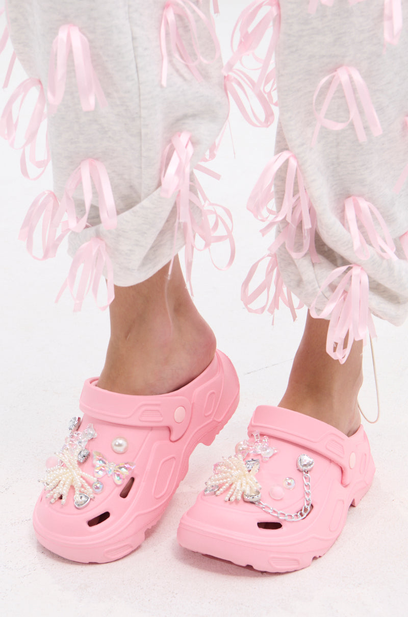 A person wears the AZALEA WANG MAVA PINK PEARL IRIDESCENT FLAT, styled with light gray pants featuring pink ribbon bows. The white background highlights the playful pearl embellishments and unique charm of their footwear.
