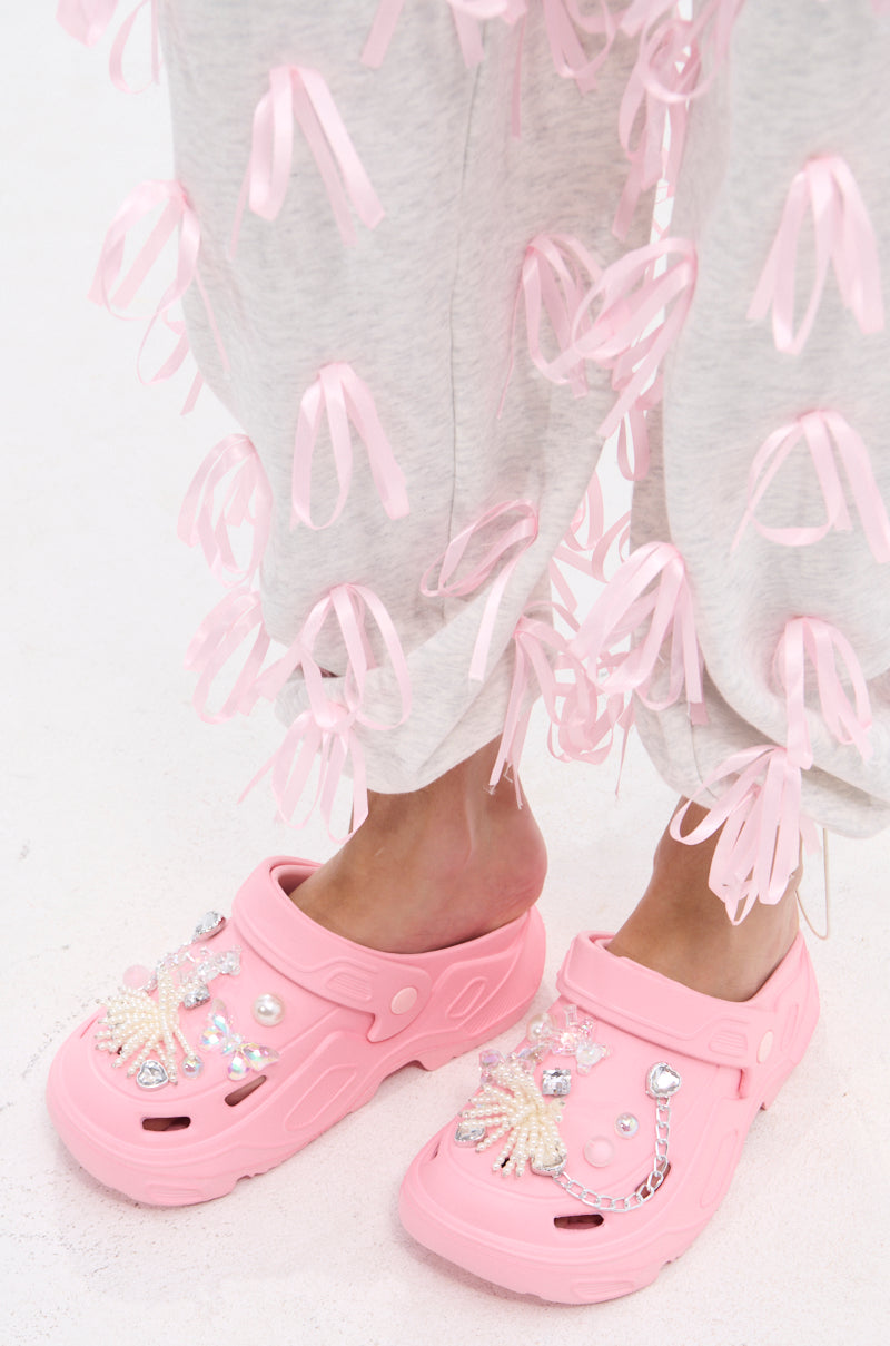Person wearing light gray pants with pink ribbon loops and the AZALEA WANG MAVA PINK PEARL IRIDESCENT FLAT, featuring beaded, jeweled, and pearl accents, standing on a light-colored surface.
