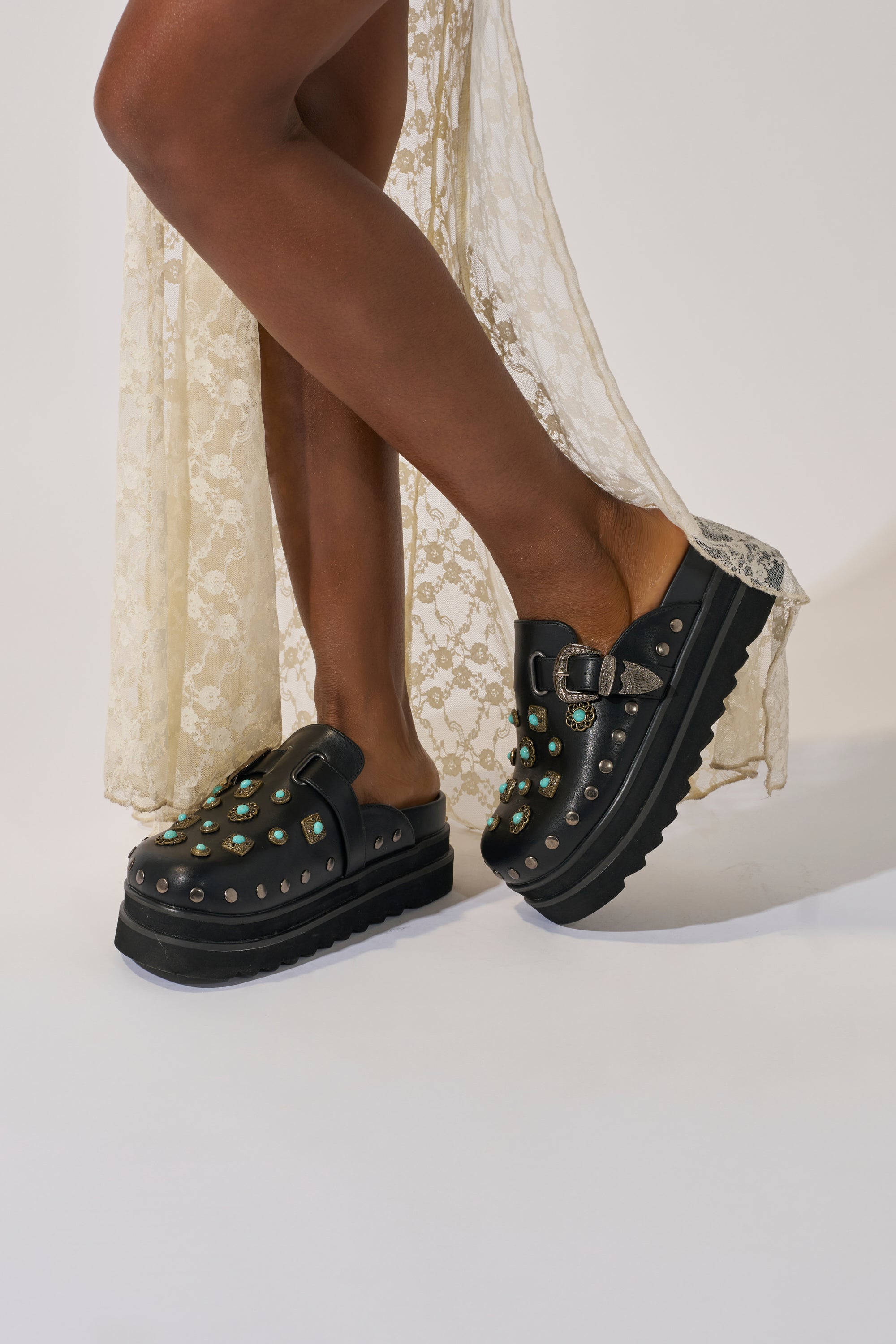 A person poses with crossed legs against a plain background, wearing a cream lace dress and the AZALEA WANG MAVIS FLAT EMBELLISHED CLOG featuring turquoise stones, silver studs, and a western buckle.