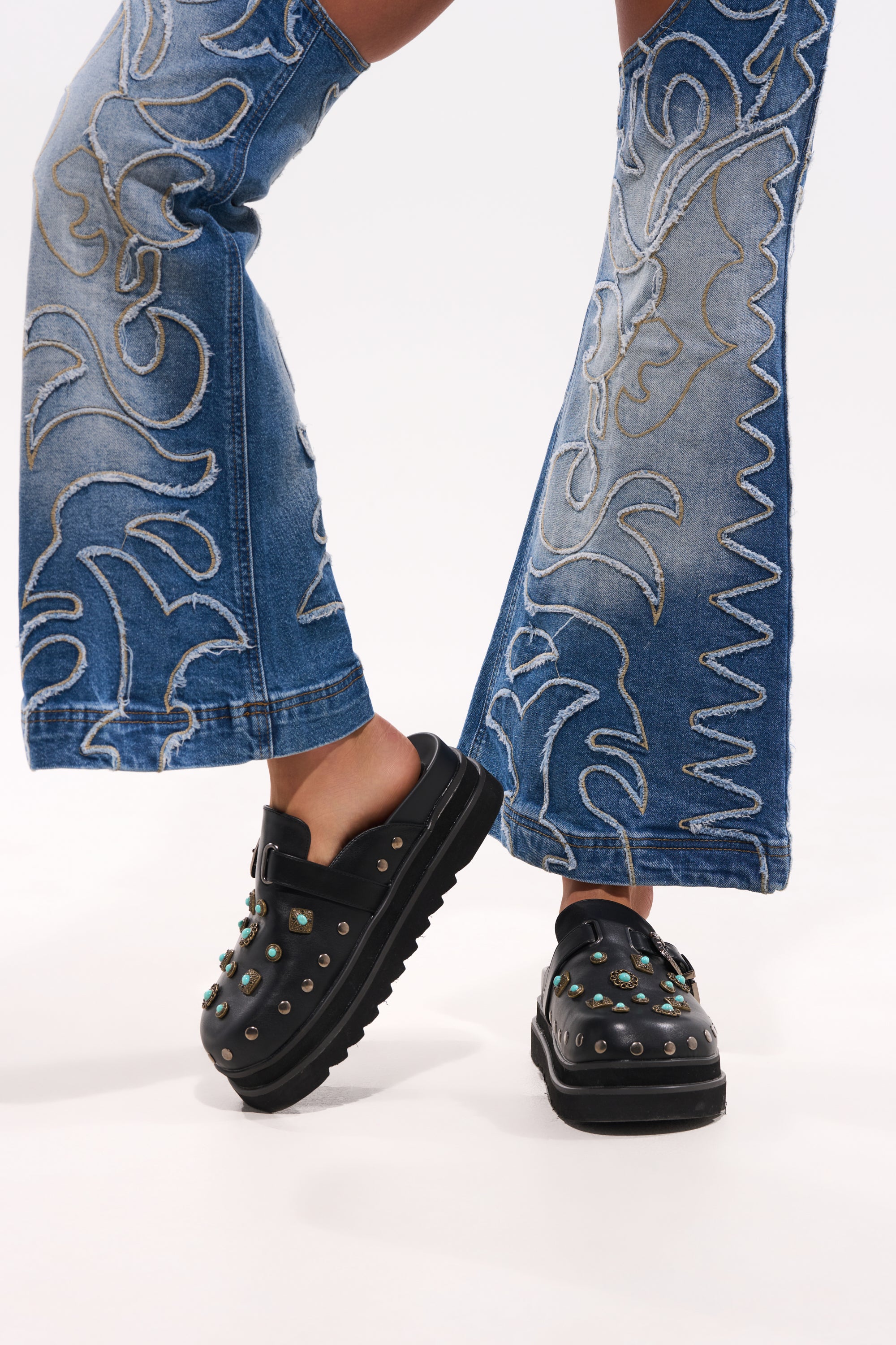 A person wears flared blue jeans with decorative stitching and the AZALEA WANG MAVIS FLAT EMBELLISHED CLOG, featuring black material and metallic stud accents, standing against a plain white background. 0