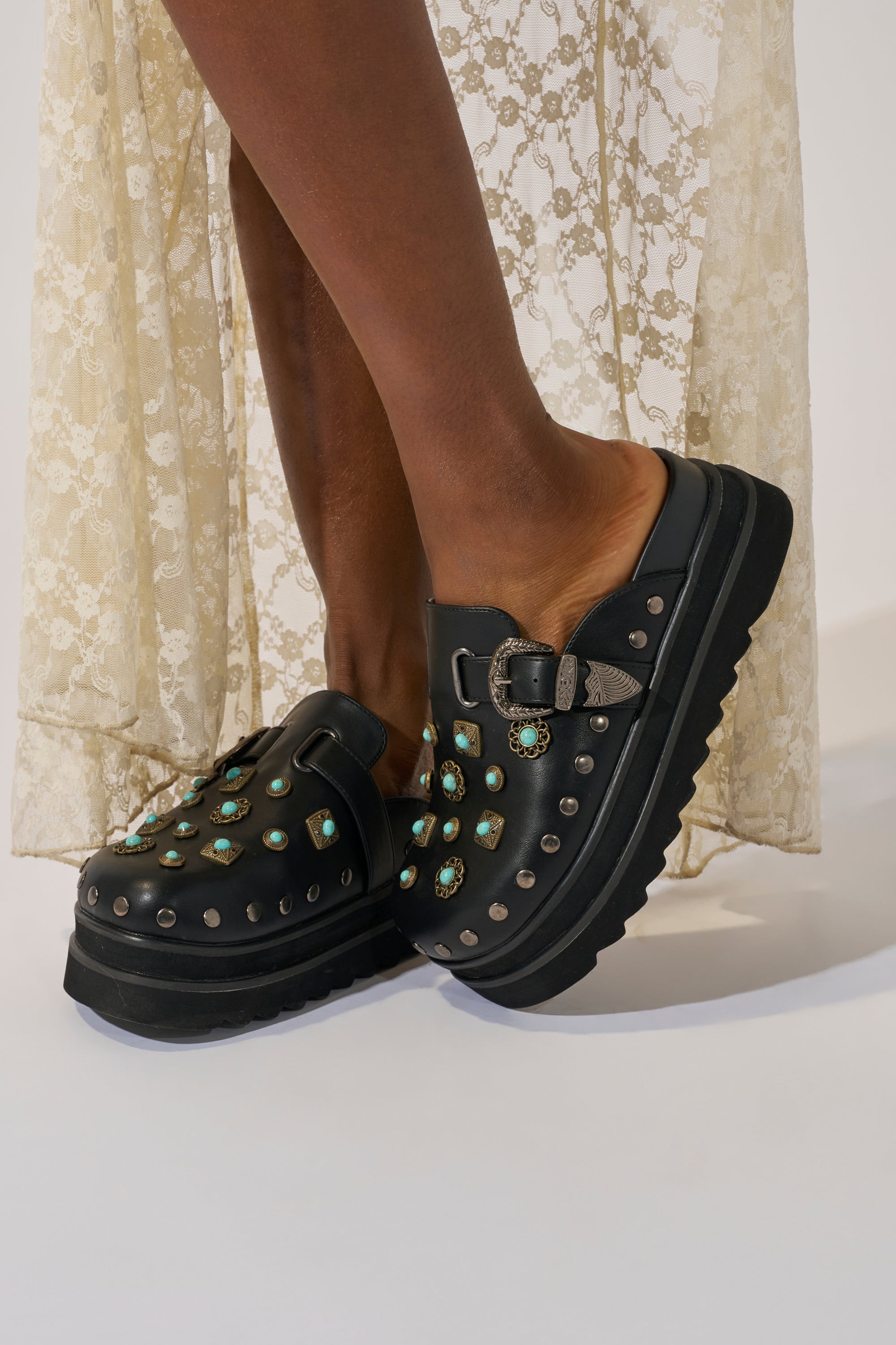 A person wears AZALEA WANG MAVIS FLAT EMBELLISHED CLOGS—black faux leather slides with turquoise and silver studs—with a cream lace dress. Only the lower legs and feet are visible.