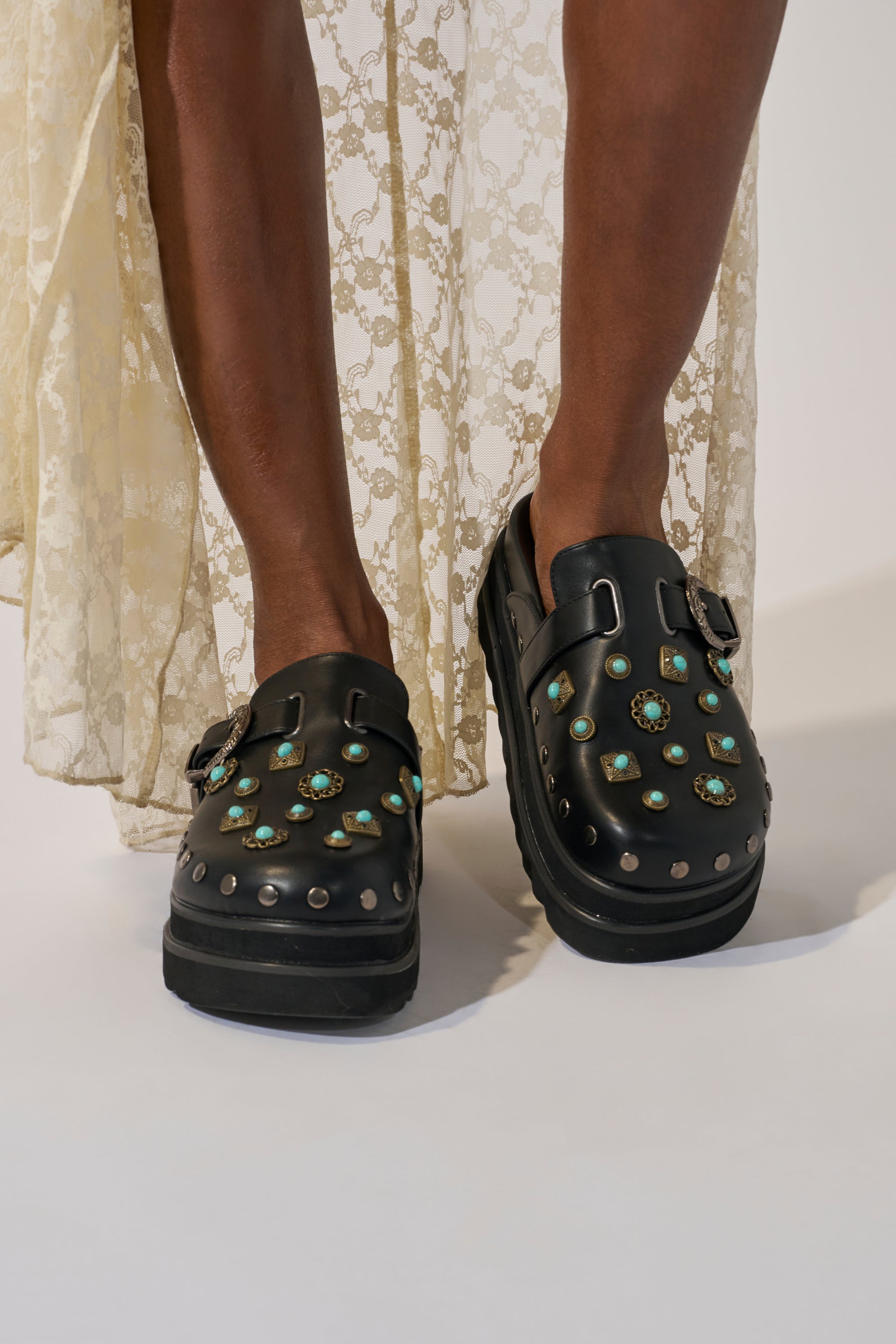 A person wearing a cream lace skirt and the AZALEA WANG MAVIS FLAT EMBELLISHED CLOG with turquoise stones and metal studs stands on a light background.