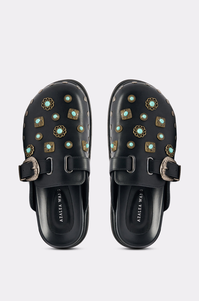 The AZALEA WANG MAVIS FLAT EMBELLISHED CLOG features black faux leather, a western buckle, and gold and turquoise-colored studs, shown from above on a light background.