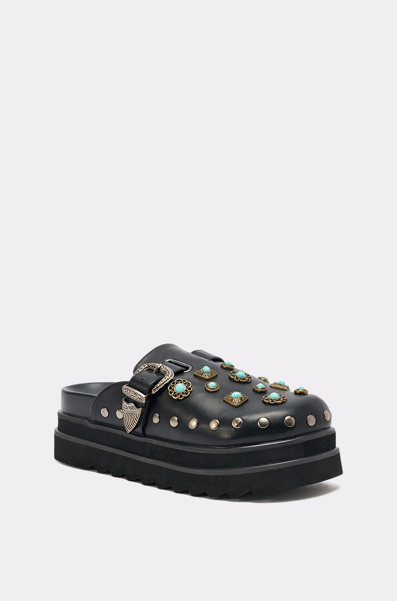 The AZALEA WANG MAVIS FLAT EMBELLISHED CLOG is a black faux leather slide platform with silver studs, turquoise floral accents, and a western buckle strap, displayed on a light gray background.