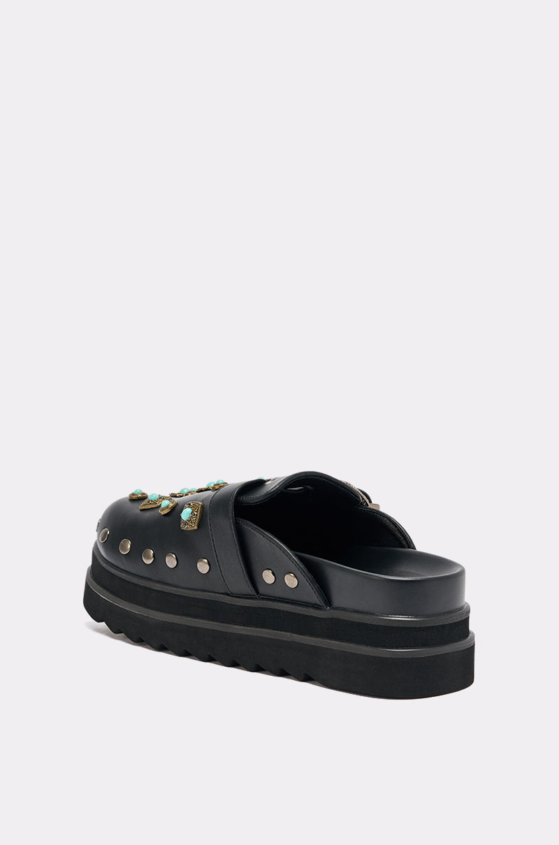 The AZALEA WANG MAVIS FLAT EMBELLISHED CLOG is a black faux leather slide sandal with silver studs and green jewel details, shown from the back at an angle against a plain light background.
