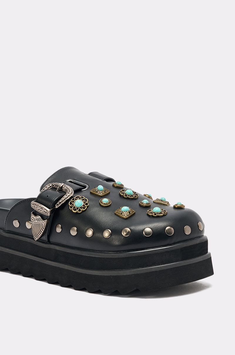 The AZALEA WANG MAVIS FLAT EMBELLISHED CLOG is a black faux leather slide featuring a western buckle, silver studs, and turquoise-accented decorations on the upper, displayed against a light background.