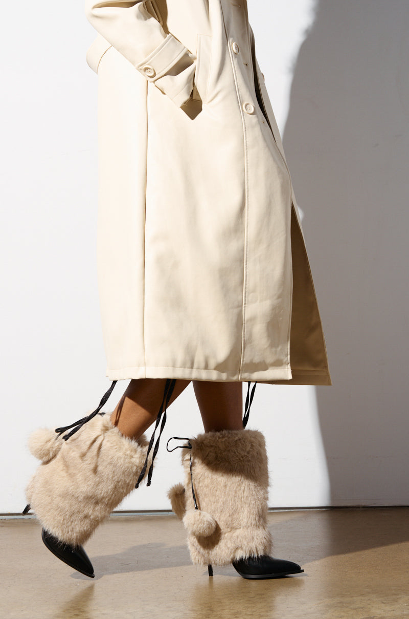 A person wearing a long beige coat and furry beige leg warmers with pom-pom details stands against a white background. Their hands are nestled in the coat pockets, while the AZALEA WANG MAYRA TAUPE BOOTIE stiletto heels elevate the look, adding a touch of sophistication to the ensemble.