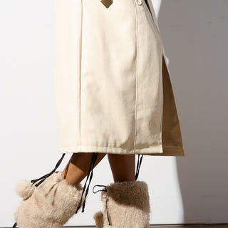 A person wearing a long beige coat and furry beige leg warmers with pom-pom details stands against a white background. Their hands are nestled in the coat pockets, while the AZALEA WANG MAYRA TAUPE BOOTIE stiletto heels elevate the look, adding a touch of sophistication to the ensemble.