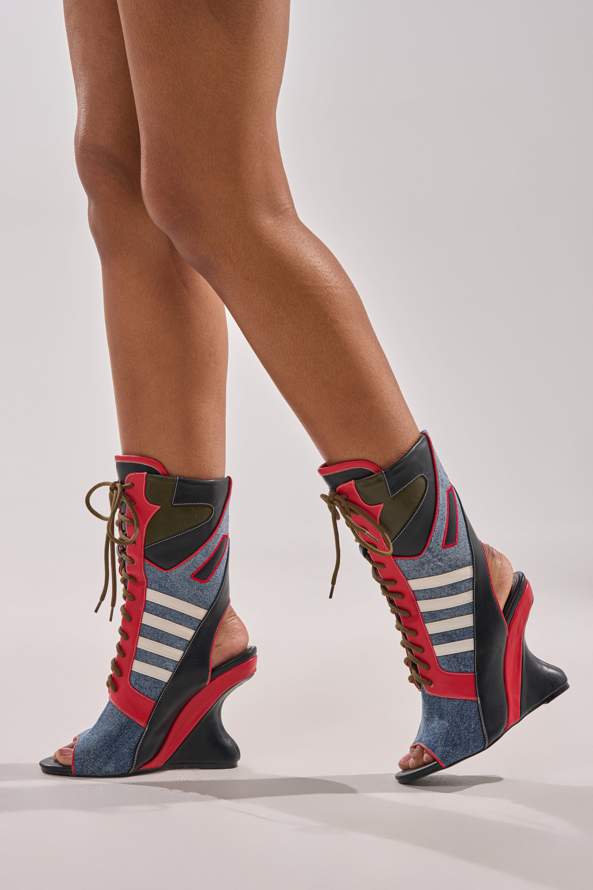 Wearing the AKIRA GOLD MAYWEATHER MULTI SPORTY LOOK WEDGE BOOTIE, featuring red, blue, black, and olive green panels with white stripes and curved heels, a person walks on a light gray surface. Only their legs and feet are visible.