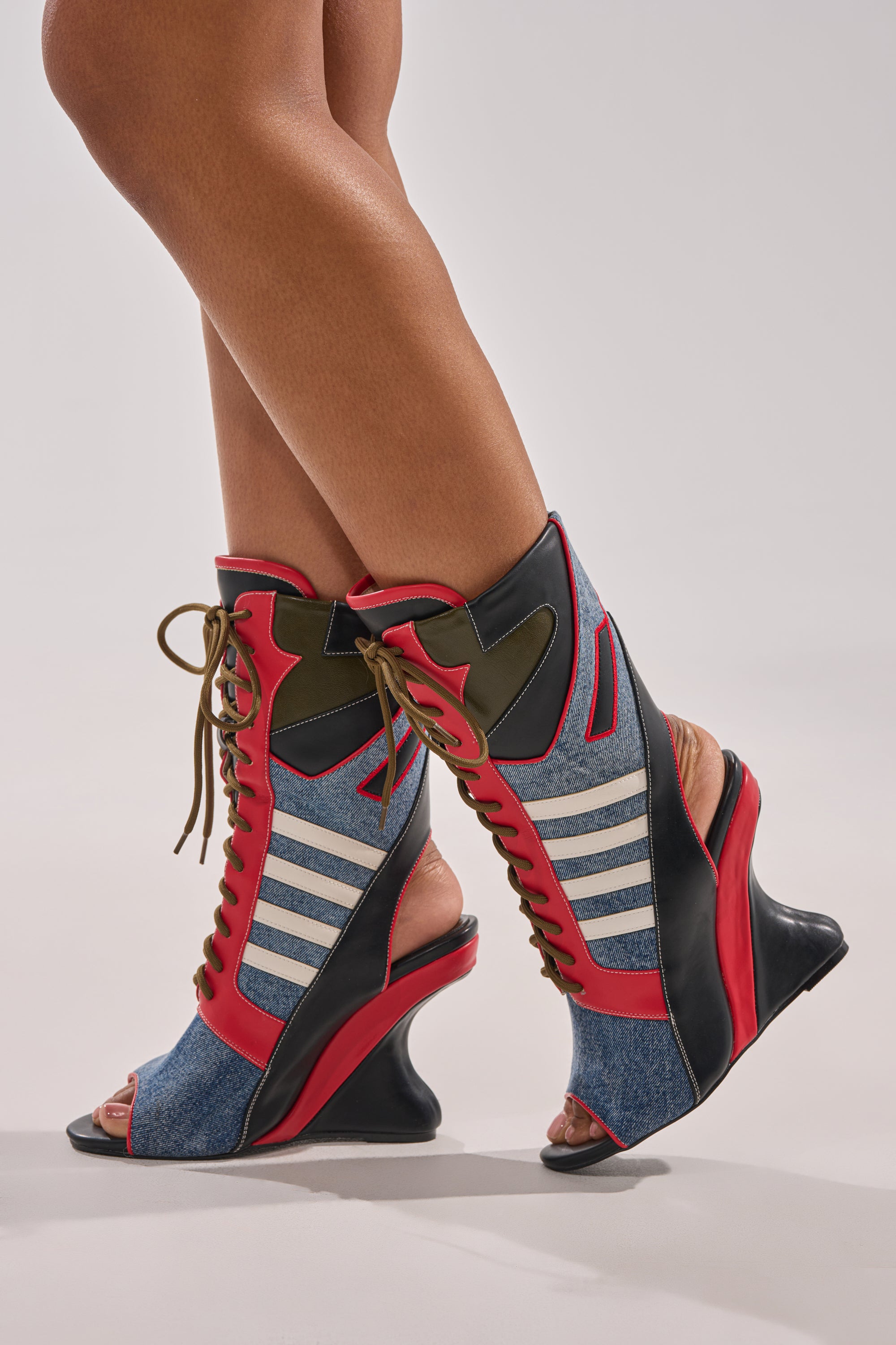 A person wears the AKIRA GOLD MAYWEATHER MULTI SPORTY LOOK WEDGE BOOTIE, featuring blue denim panels, red and black accents, gold laces, white stripes, and open toes and heels, standing with legs crossed against a neutral background.