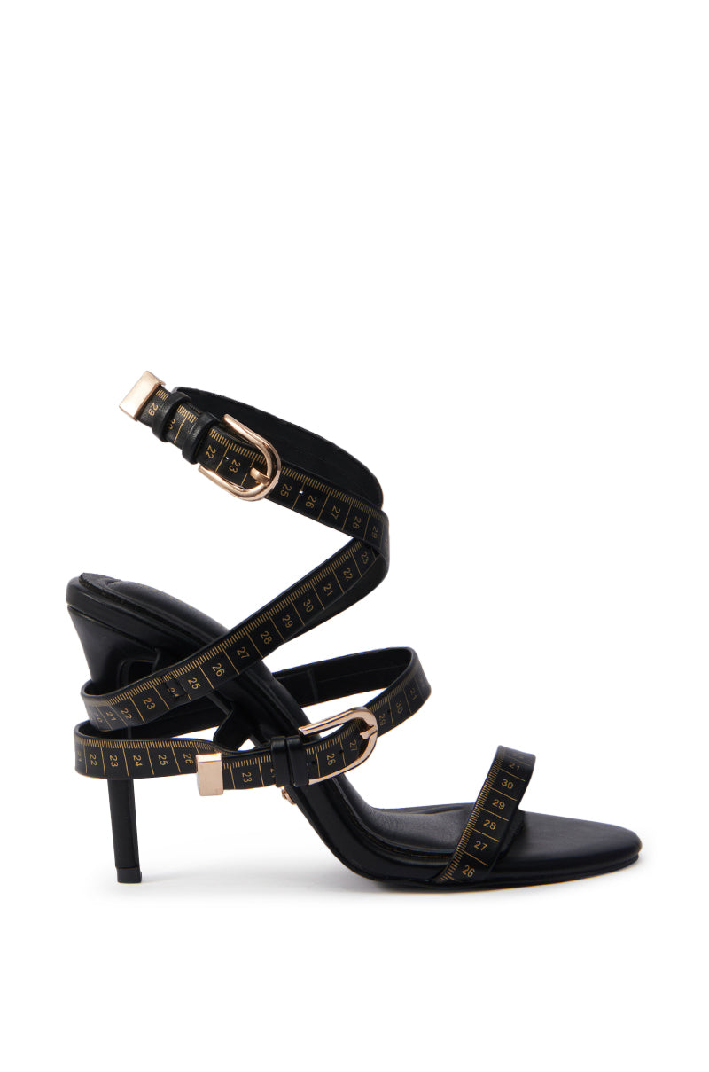 The AZALEA WANG MAZOLE BLACK TAPE MEASURE DETAIL WRAP UP SANDAL features stiletto heels, gold buckles, and faux leather measuring tape straps that wrap elegantly around the ankle and foot.