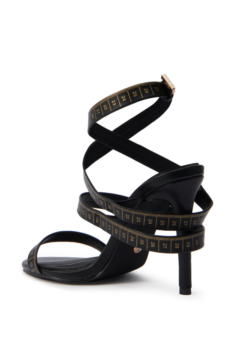 The AZALEA WANG MAZOLE BLACK TAPE MEASURE DETAIL WRAP UP SANDAL is a black high-heeled faux leather sandal with wraparound ankle straps styled to resemble a yellow and black measuring tape.